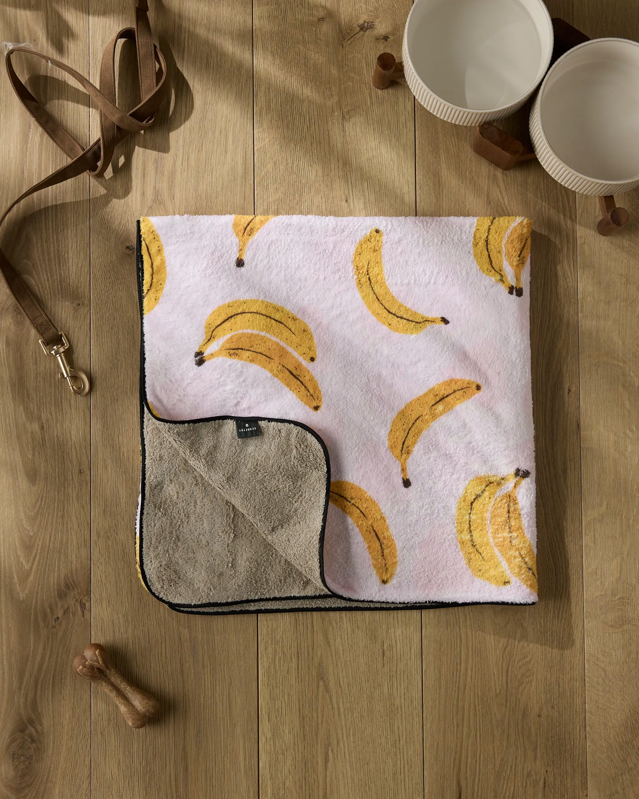 The Geometry Pupnana is a plush dog towel featuring a banana pattern on one side and plain brown on the other, shown laid out on a wooden floor with two dog bowls, a leather leash, and a wooden dog toy nearby.