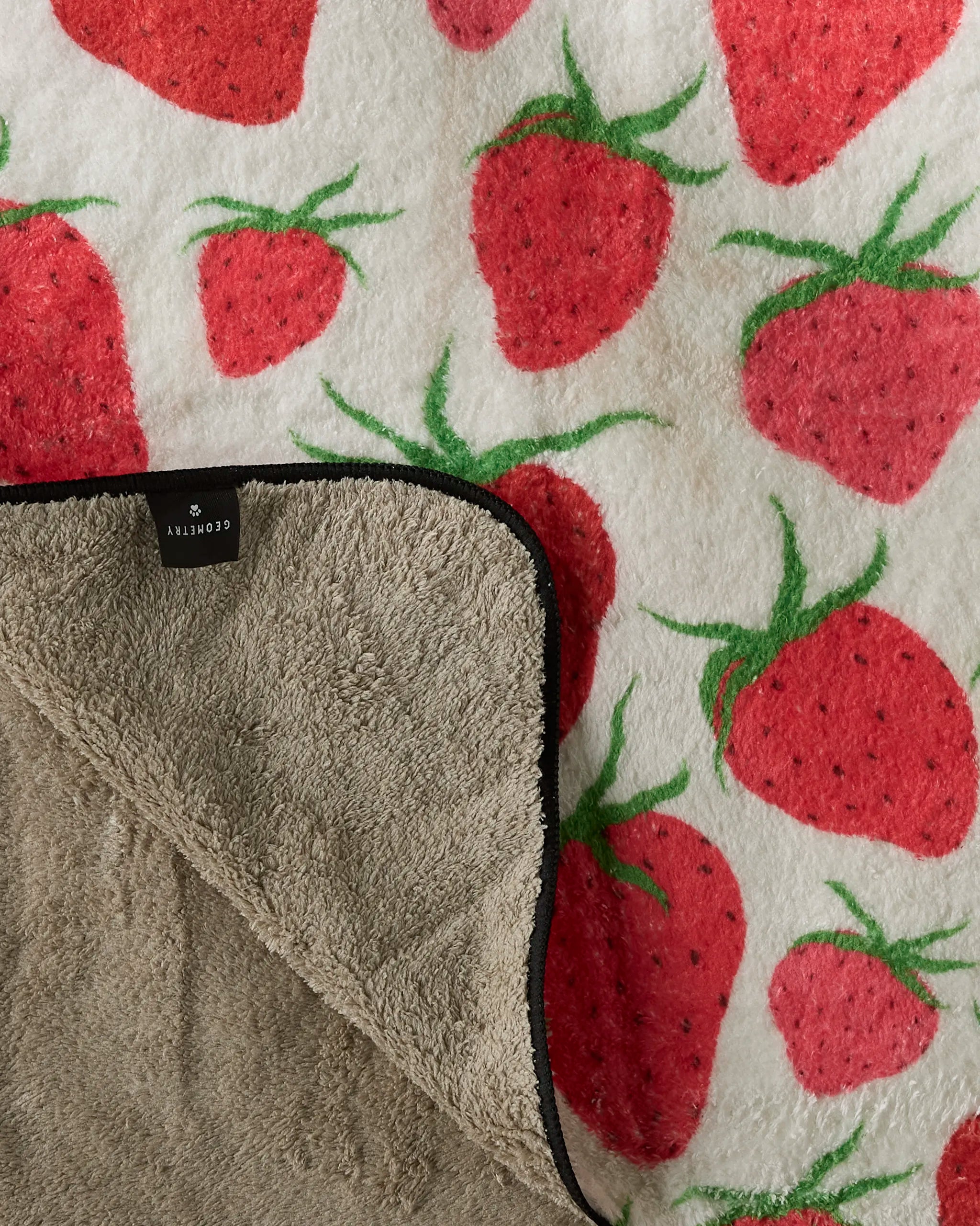 The Geometry Strawberry Sniff is a soft, ultra-absorbent dog towel featuring a vibrant strawberry pattern on white, a plain beige underside, and a small black tag along the edge.