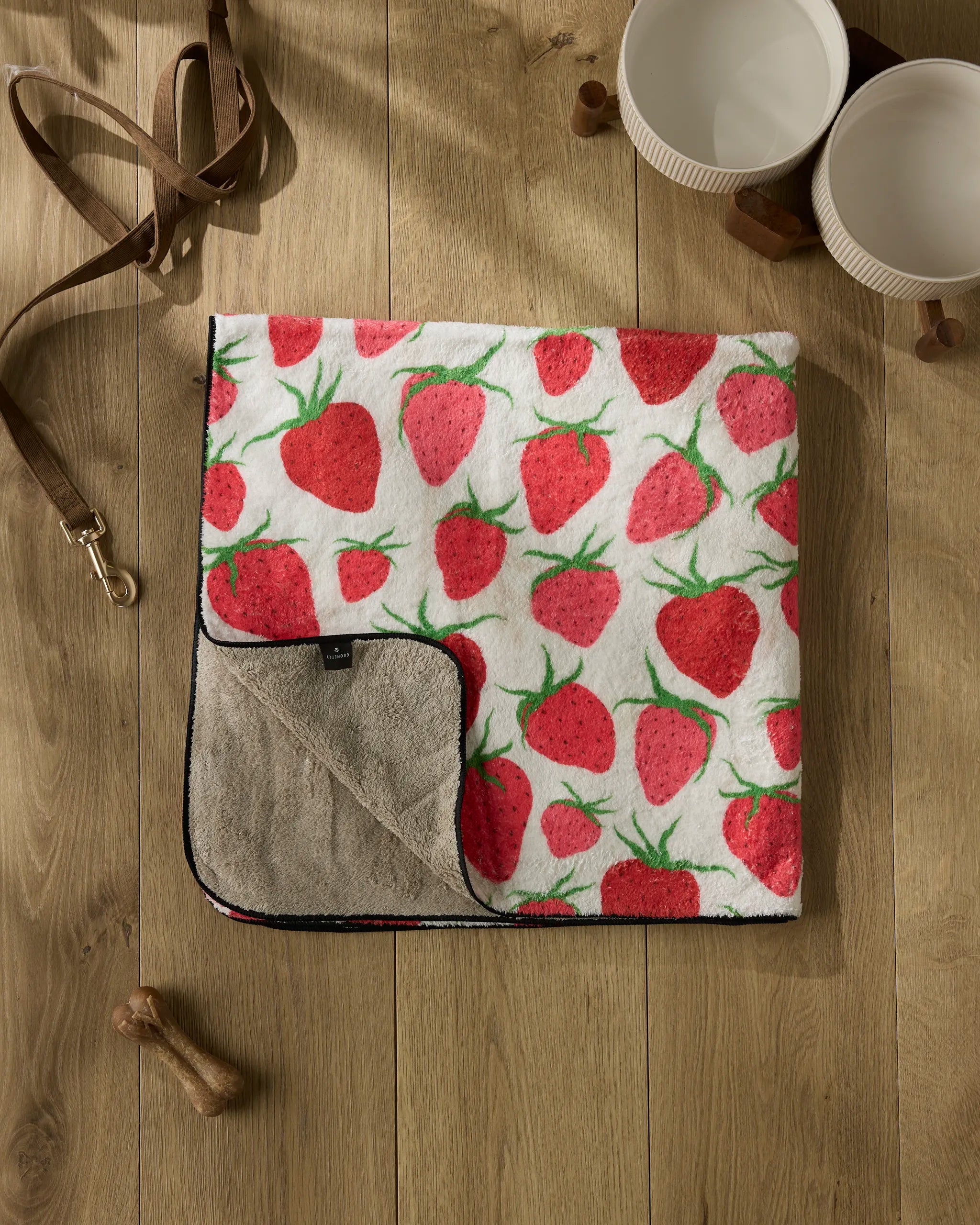 The Geometry Strawberry Sniff plush dog towel, featuring a red strawberry pattern, is displayed on a wooden floor alongside a dog leash, two ceramic bowls, and a small dog bone toy.