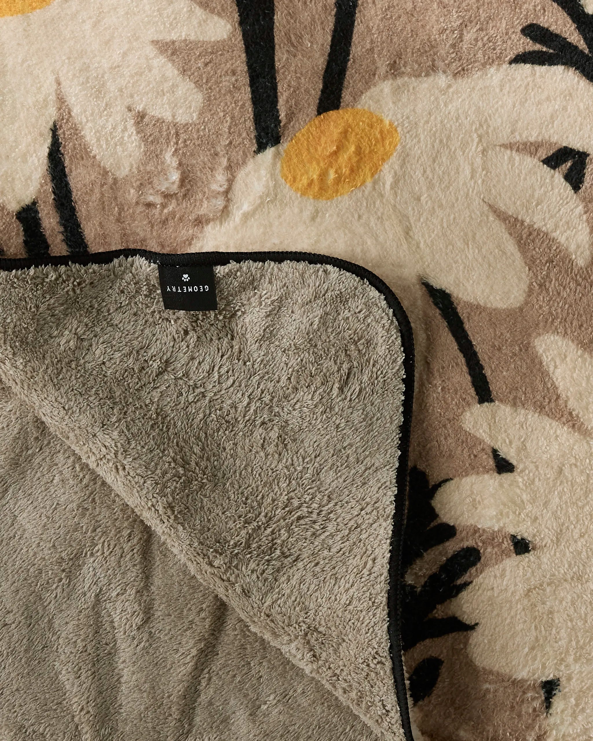 A close-up of the Geometry Sweet Daisies rug shows its fluffy beige texture with white and yellow daisy patterns. A folded corner reveals the solid gray underside and a black label with white text.
