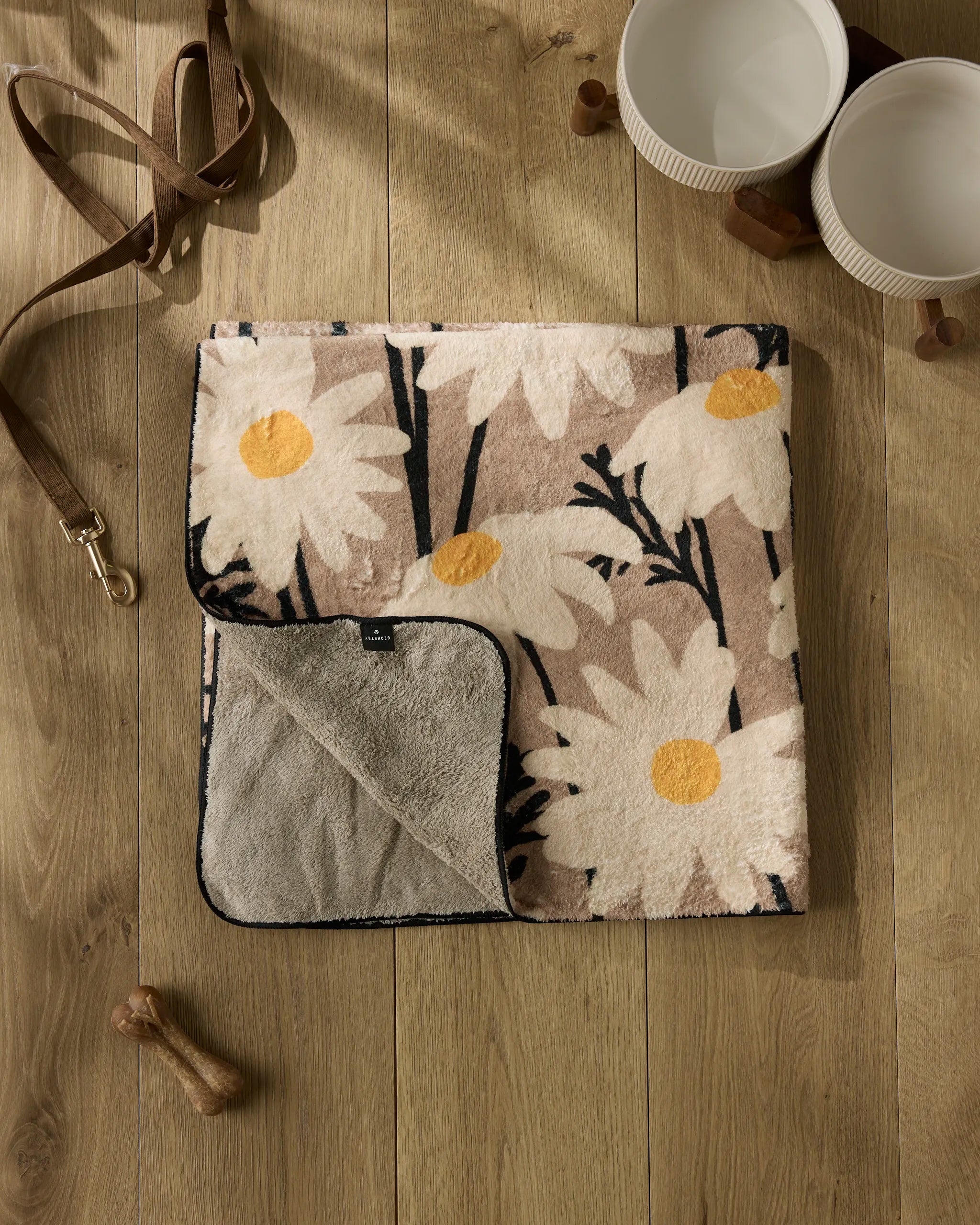 The Geometry "Sweet Daisies" plush dog towel with a daisy pattern and gray backing is folded on a wooden floor next to two white bowls, a brown leash, and a small wooden bone toy.