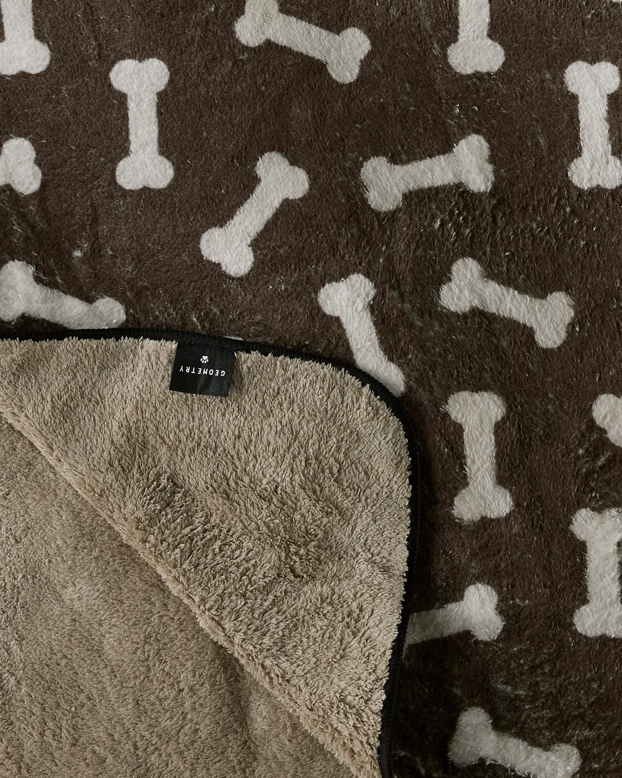 A soft brown plush dog towel from Geometry's Sweet Treats collection, featuring white bone patterns, a fluffy beige underside, and a small black SORVITY tag.