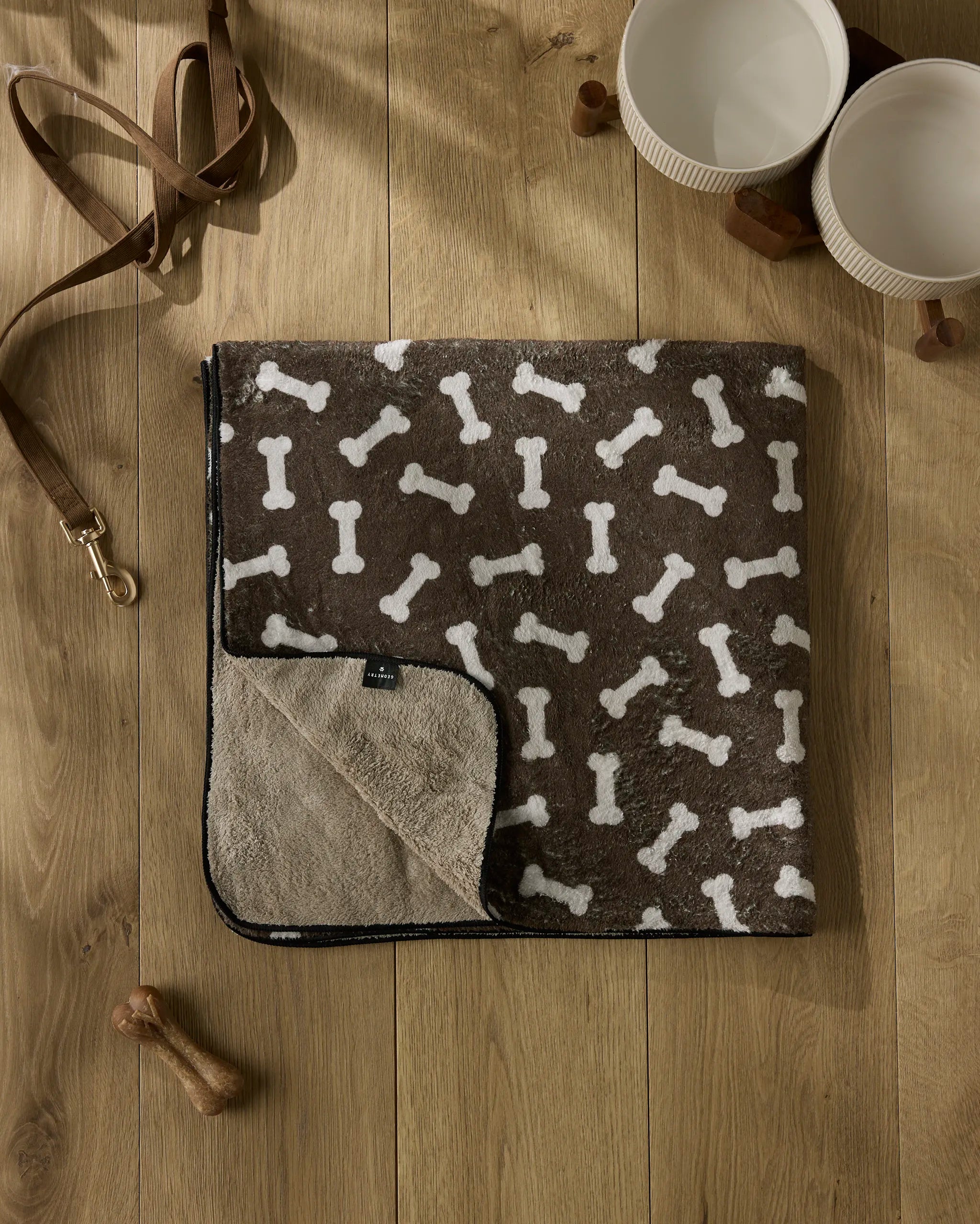 The Geometry "Sweet Treats" plush dog towel in brown with a white bone pattern lies on a wooden floor, next to two white bowls, a brown leash, a dog bone toy, and two wooden cubes.