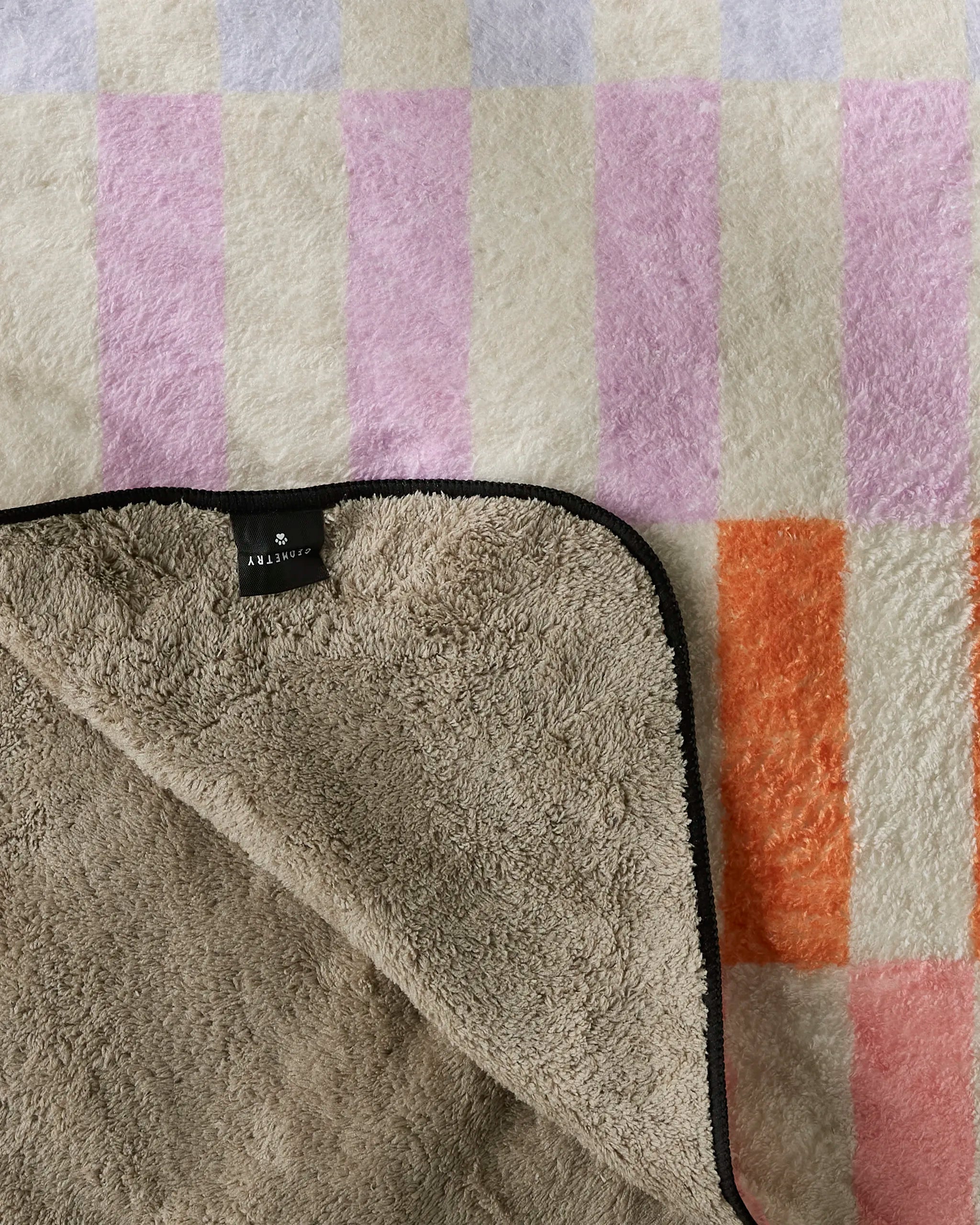 The Geometry Wag Wise plush dog towel features a pastel checkered pattern in purple, pink, and orange with a beige underside and a black label with white text. One corner is folded to show the soft lining.
