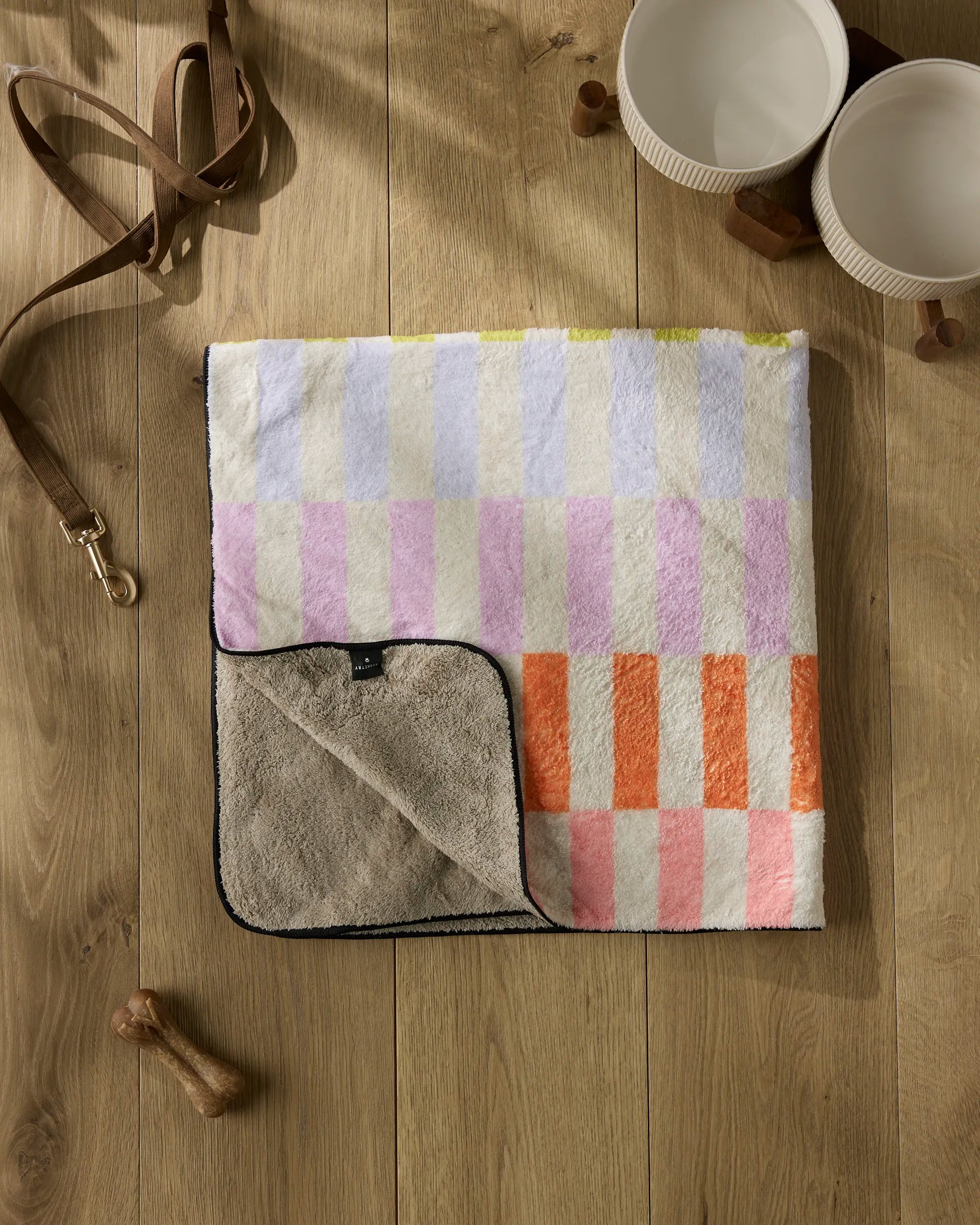 The Geometry Wag Wise ultra-absorbent dog towel, featuring soft pastel and orange squares, is folded on a wooden floor beside two white bowls, a brown leash, and a wooden bone toy.