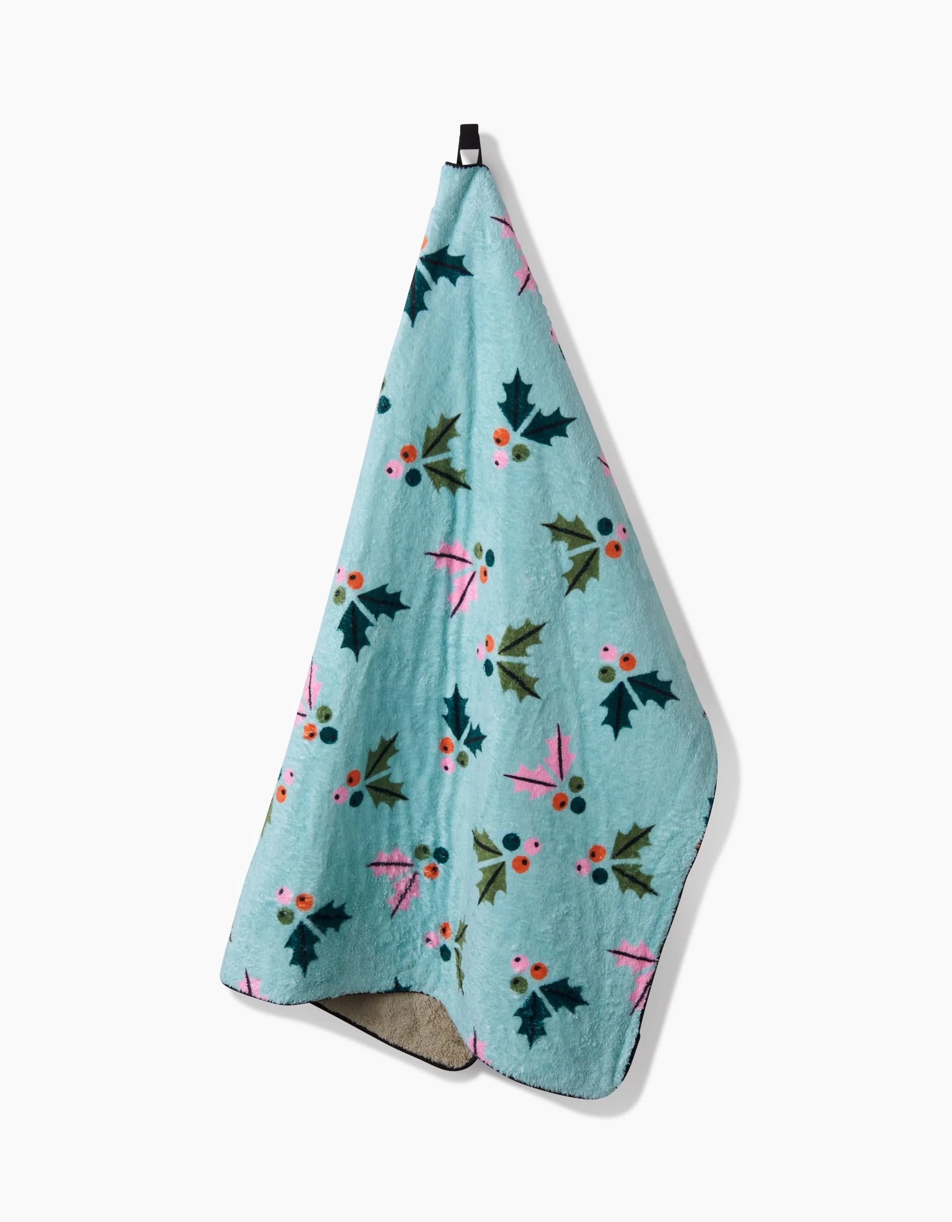 The Megan Roy Berries Mint dog towel in soft blue, decorated with festive green holly leaves and red berries, hangs from a black loop against a plain white background.