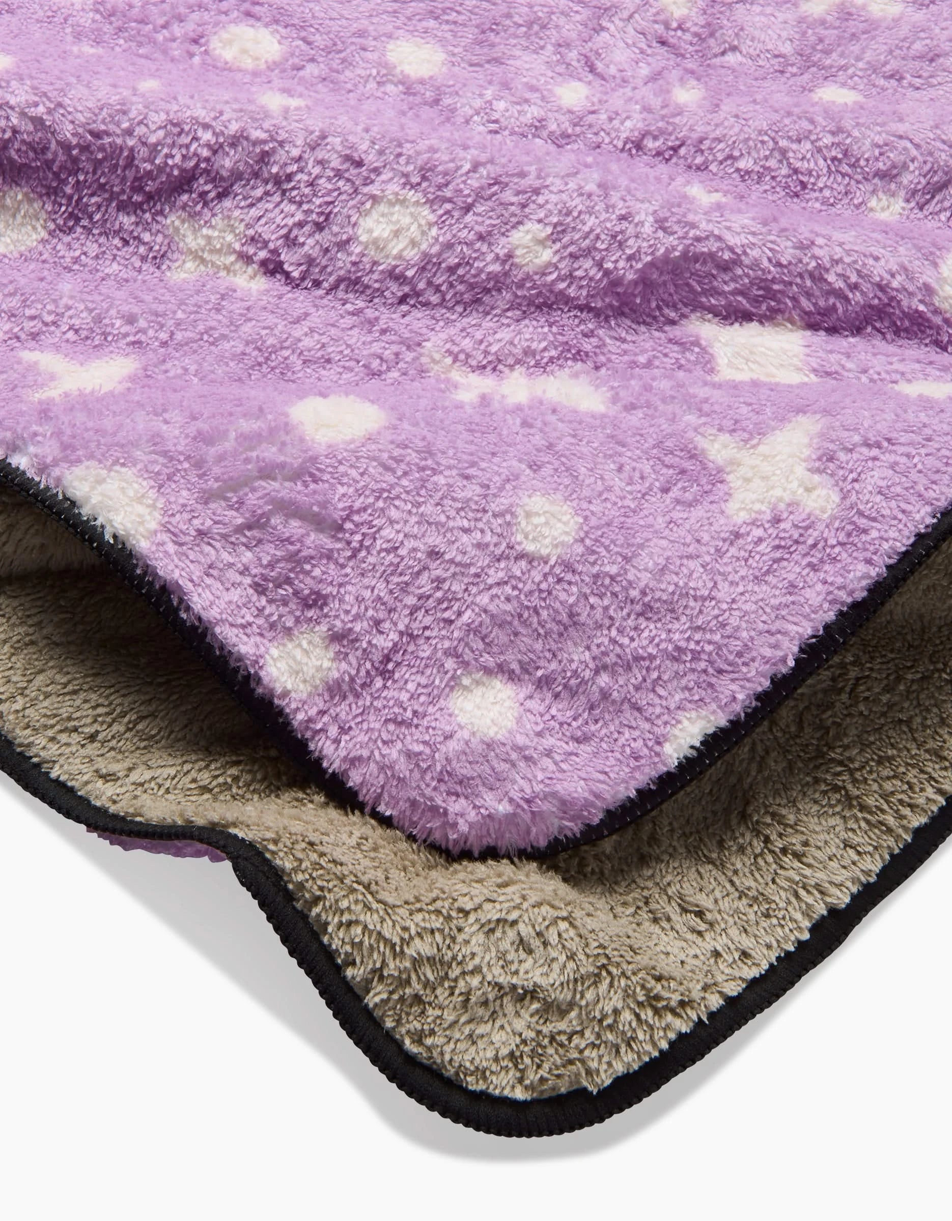 A close-up of two stacked fleece blankets: the top is Amy MacCready’s Estrella Purple with white polka dots and stars; below is a beige blanket with a black edge, ultra-absorbent for plush comfort.