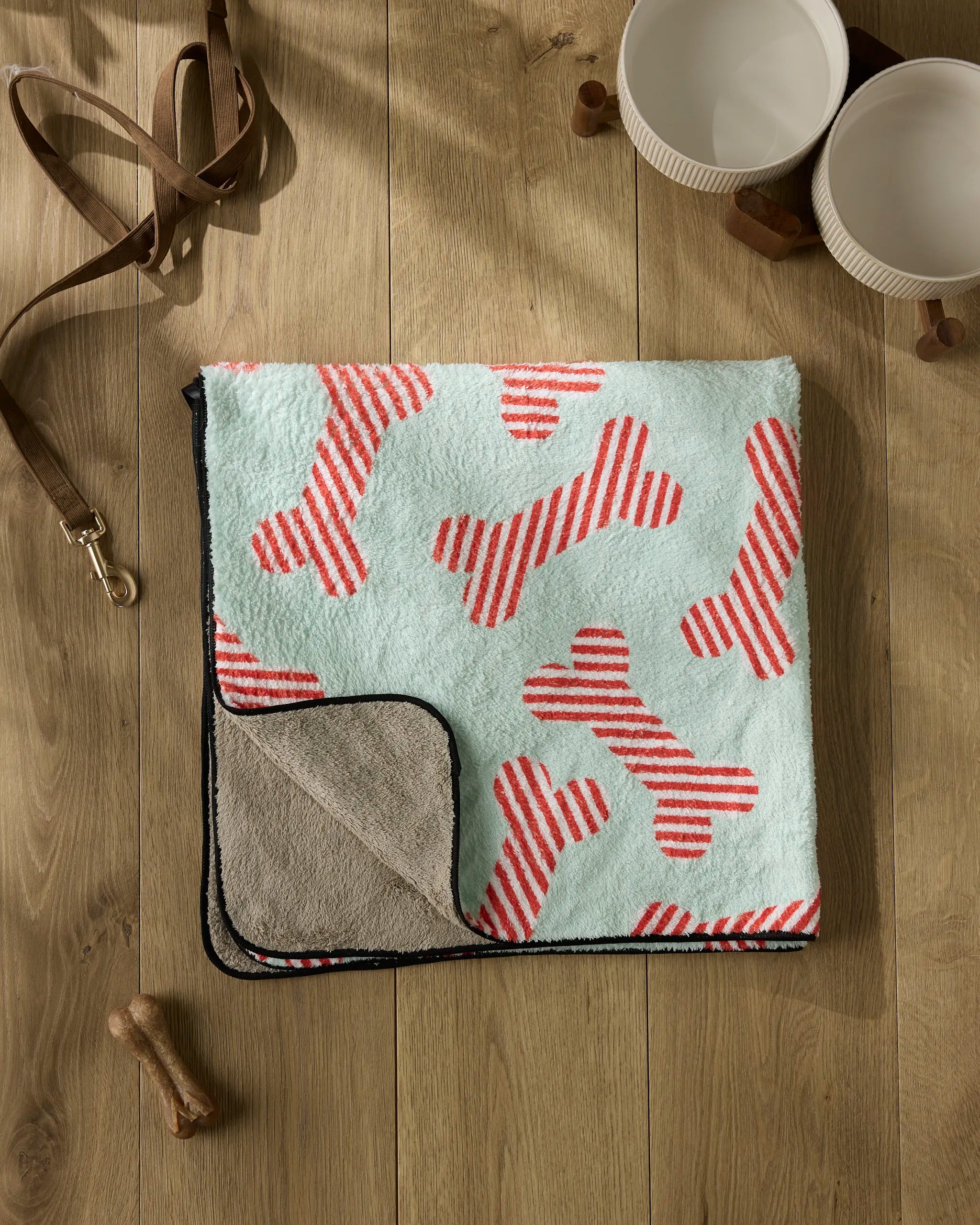 The Geometry Yuletide Pup is a folded plush dog towel featuring red and white striped bone patterns on a light background, shown on a wooden floor beside a leash, two empty pet bowls, and a chew bone.