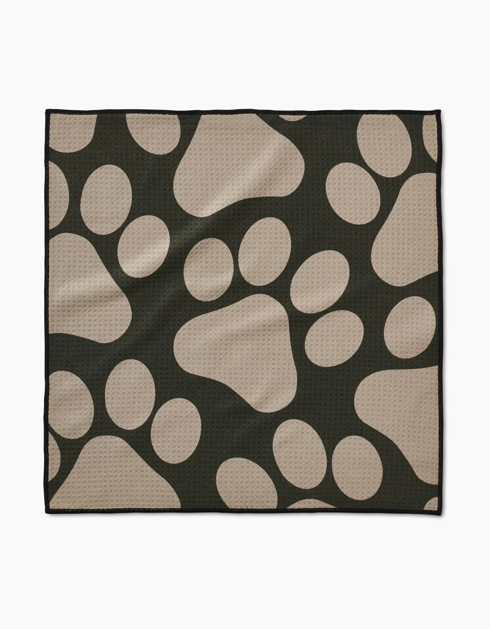 The Geometry Paw Prints mat is a black, rectangular towel with large tan paw prints on its ultra-absorbent, textured surface. Made from recycled materials, it has a clean black border for a simple finish.