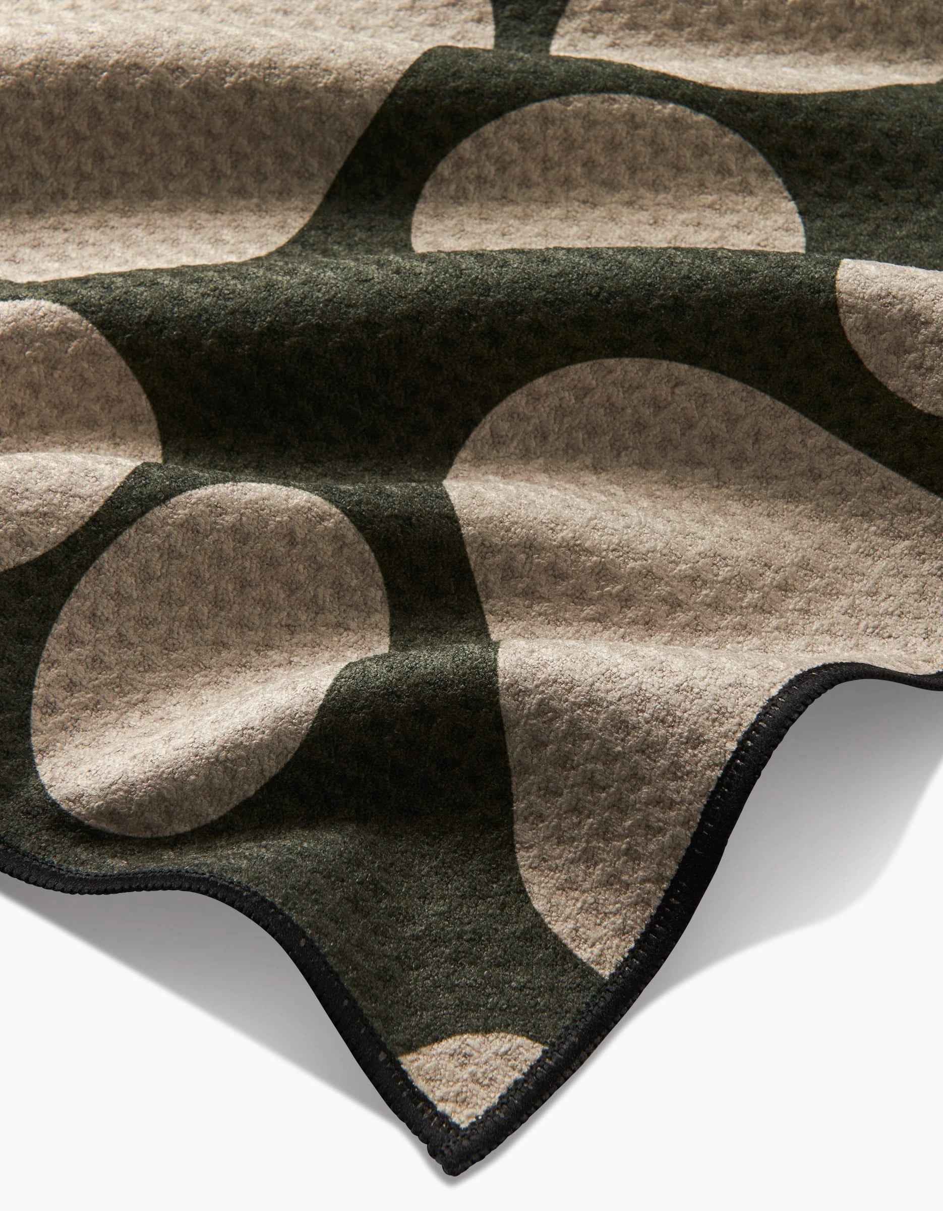 A close-up of Geometrys Paw Prints towel, crafted from recycled materials with a bold beige and dark green abstract pattern and soft texture, featuring wavy edges trimmed in black.