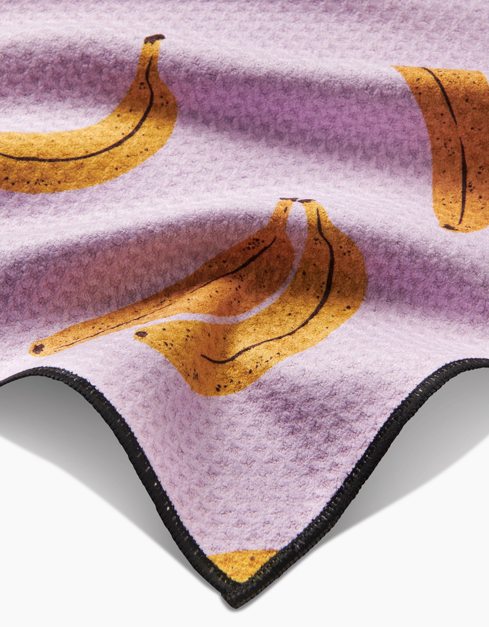 The Geometry Pupnana Paw Towel is lilac with a wavy edge, black trim, and a repeating banana print. Made from ultra-absorbent Geoweave fabric for textured drying.