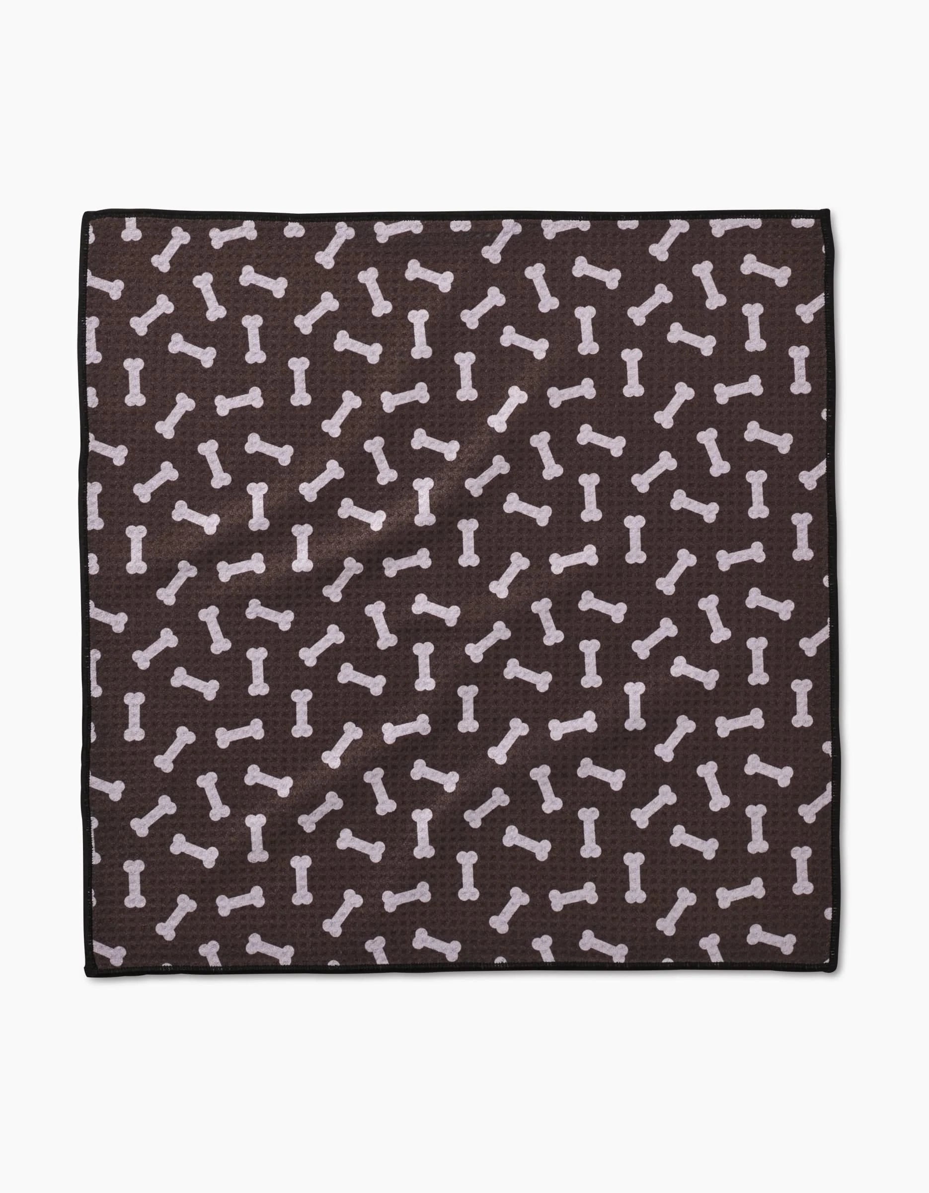 The Sweet Treats Paw Towel by Geometry is a square, dark brown towel made from recycled materials, featuring a repeating pattern of light-colored dog bones in various directions.