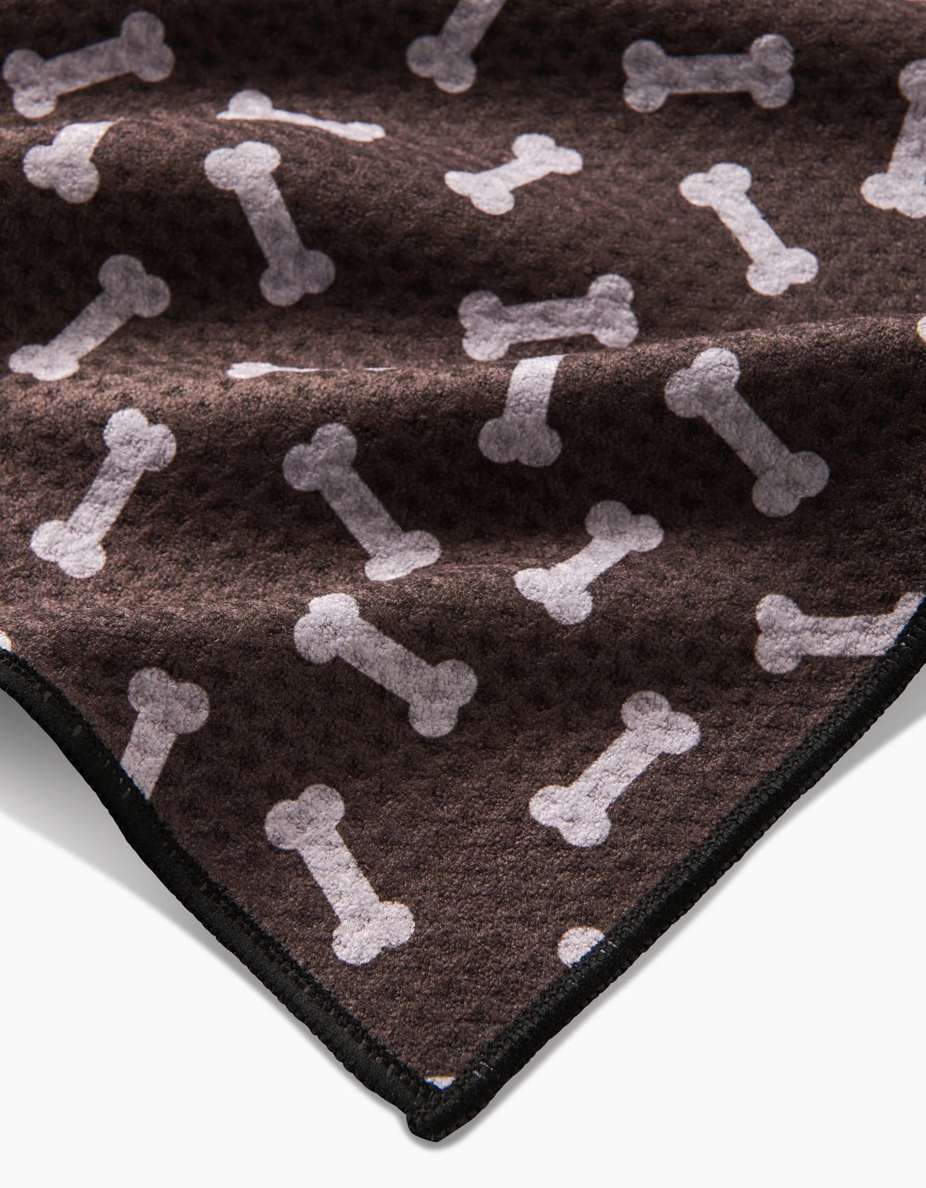 The Geometry Sweet Treats is a soft, brown fleece blanket made from recycled materials, featuring a repeating white dog bone pattern and black stitched edge, shown partially folded.