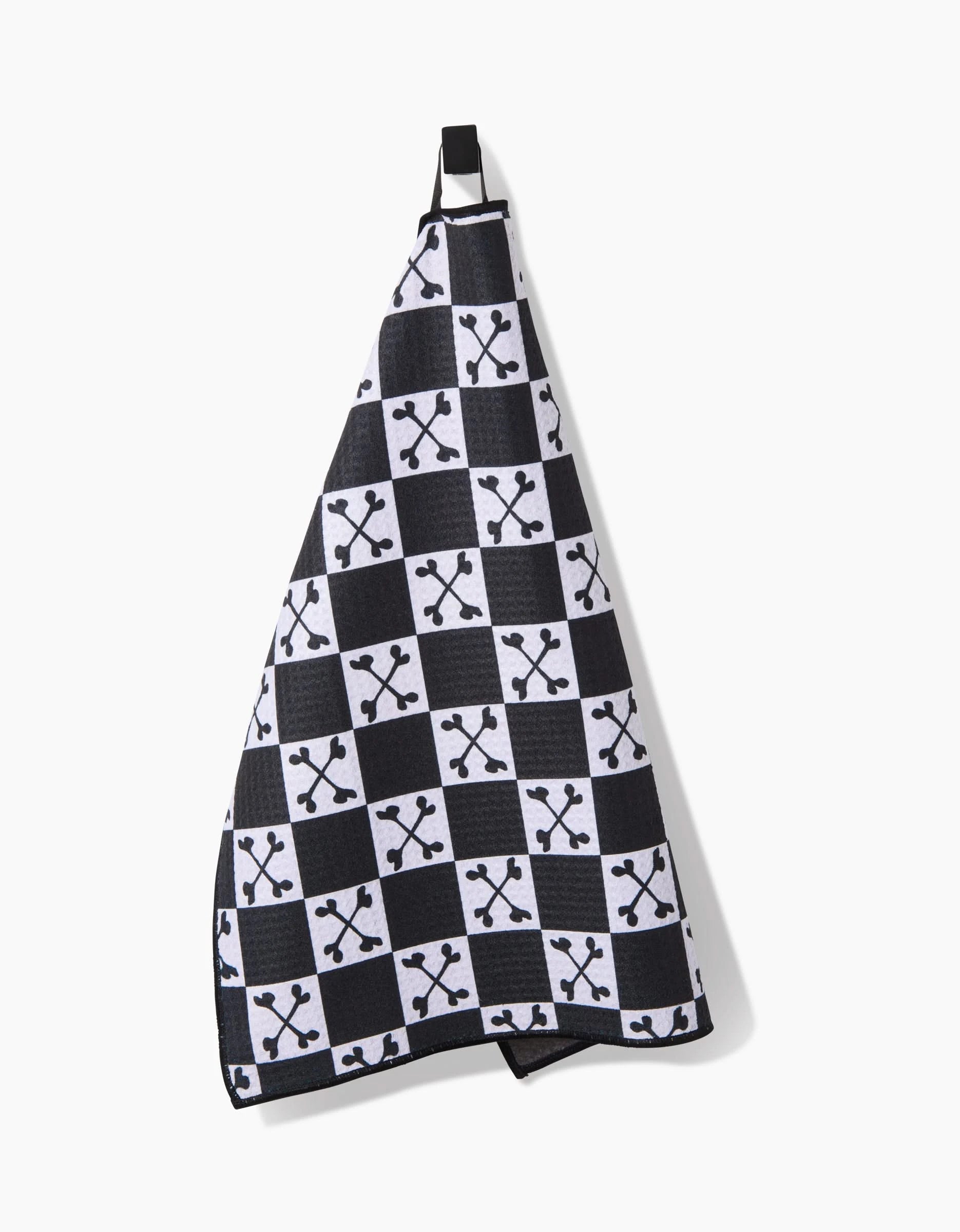 The Boneyard White Checkerboard Paw Towel by Charlie Rowan Designs hangs on a hook, showcasing a black and white checkered pattern with small crossed bones. Made from ultra-absorbent recycled materials, its stylish and eco-friendly.
