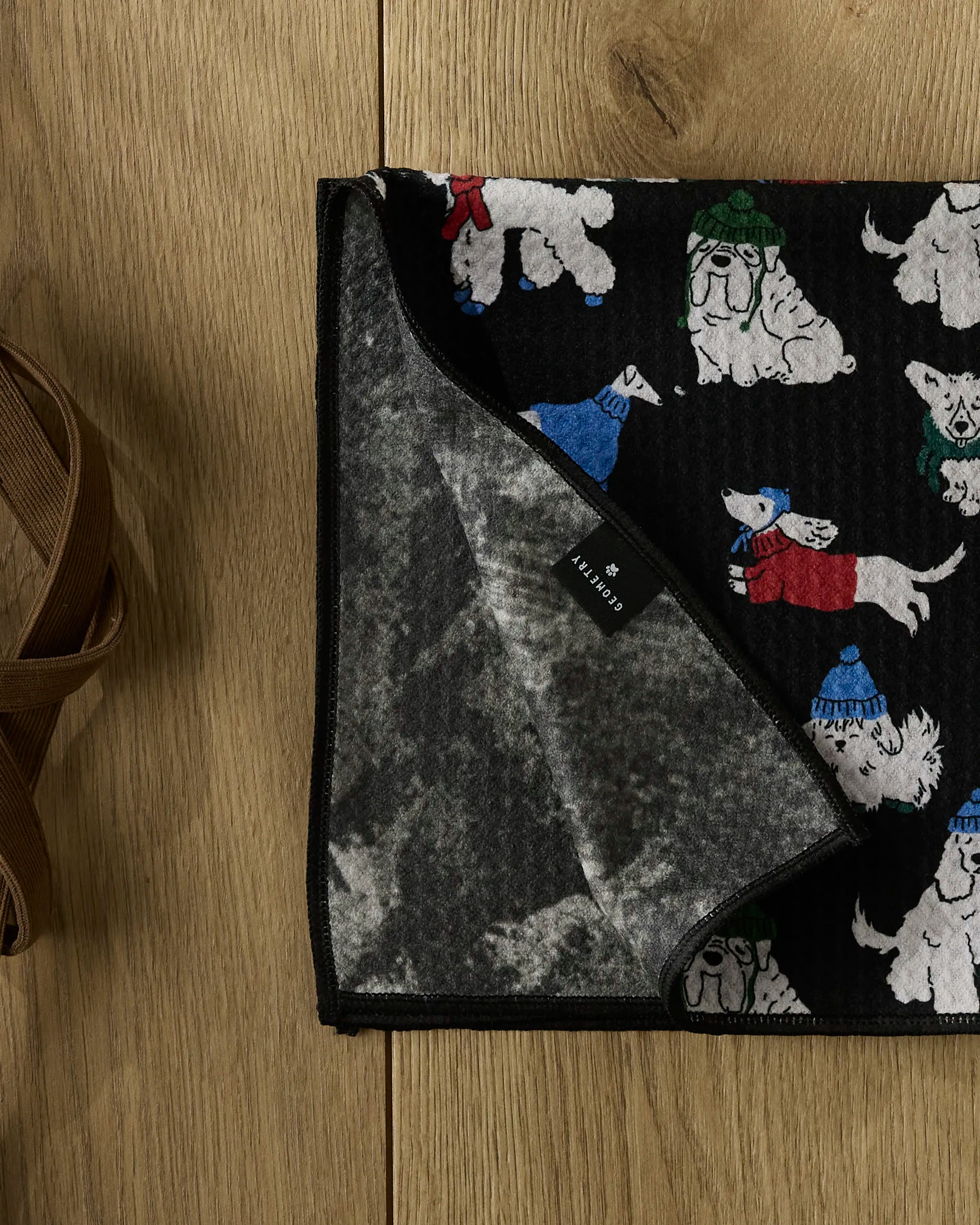 A Geometry Dogue Magazine pet towel lies on a wooden floor, featuring black fabric with illustrations of white dogs in colorful hats and scarves. The corner is folded to show its gray, textured underside and label—ultra-absorbent and cozy for your pet.