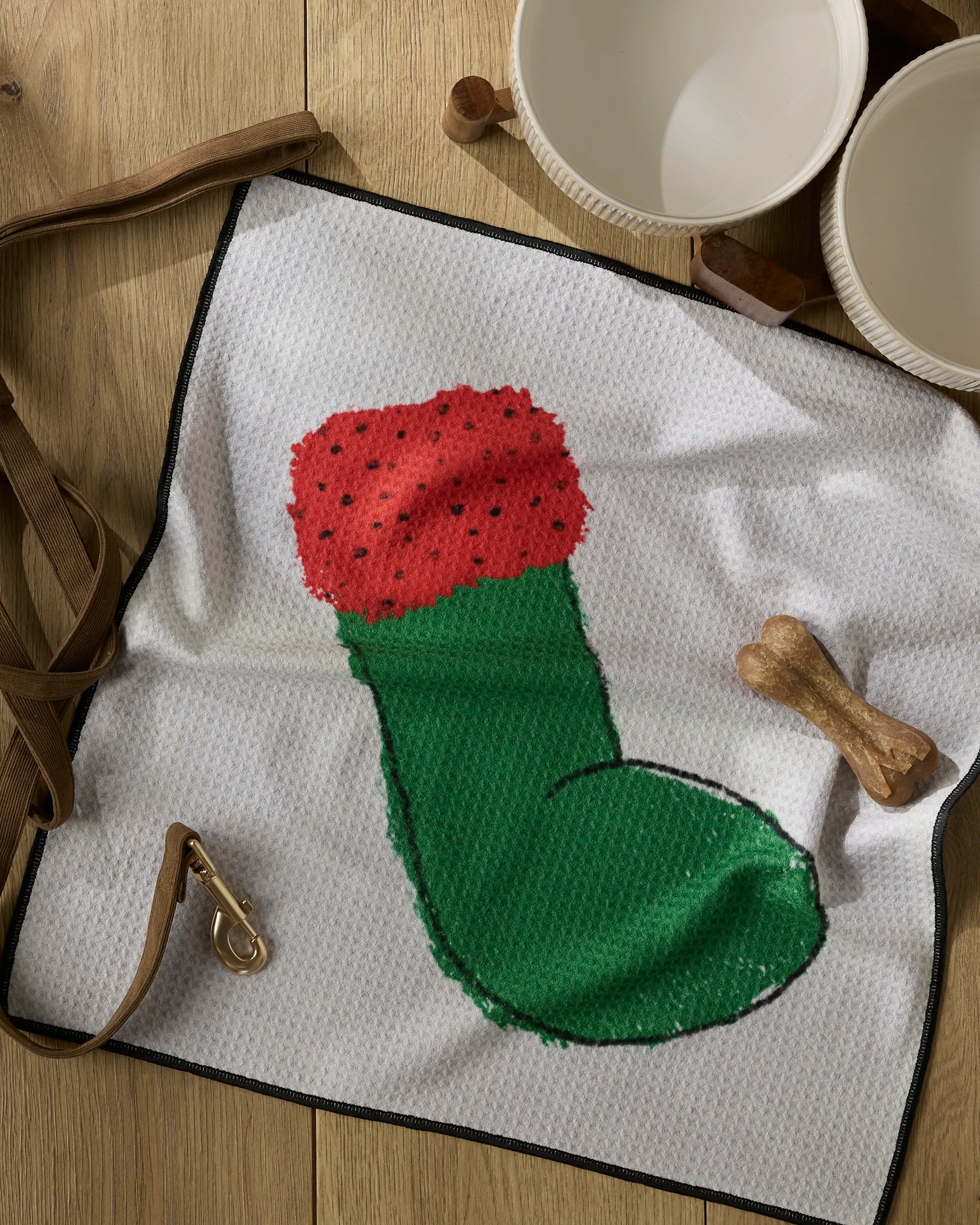 The Geometry Silly Stocking, a white paw towel made from recycled materials with a red and green Christmas stocking design, rests on a wooden surface beside a dog leash, bone-shaped treat, and white ceramic bowls.