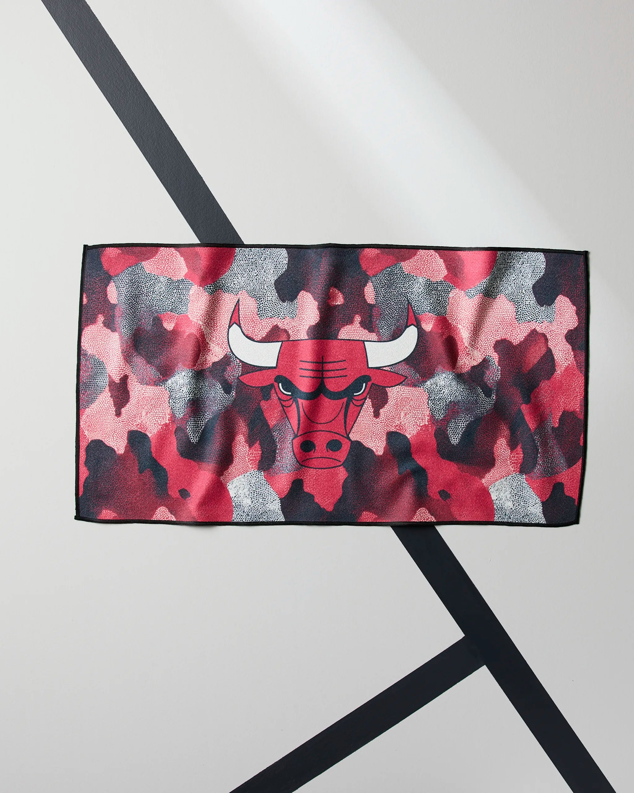 The Geometry Bulls Camo towel features a rectangular shape, a red, pink, gray, and black camouflage pattern, and a bull’s head logo in the center. It’s shown hanging on a wall with diagonal black lines.