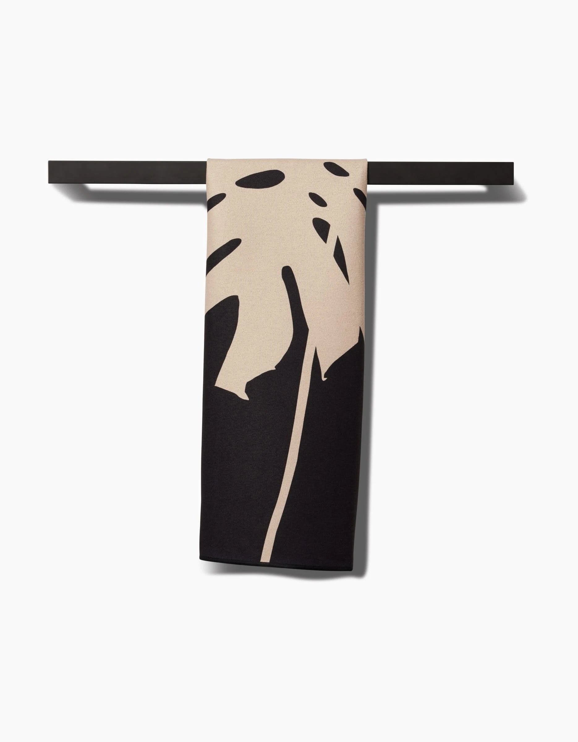 The Cream Monstera Fitness Towel by PrintedPlant, featuring a bold abstract leaf design and crafted from recycled materials, hangs neatly over a black rod against a white background.