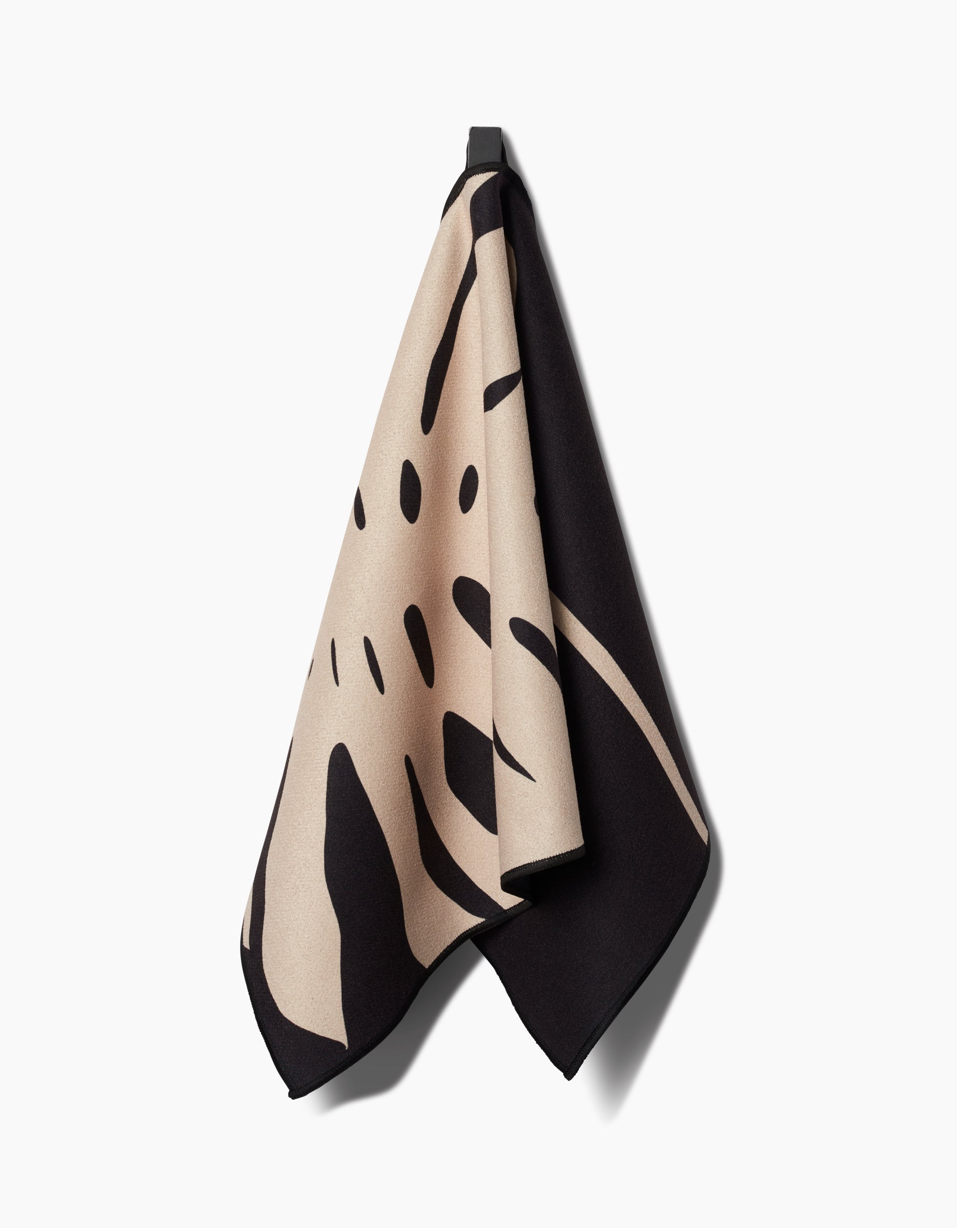 The PrintedPlant Cream Monstera Fitness Towel, featuring a beige and black leaf-inspired pattern, hangs on a hook against a white background. Made from ultra-absorbent fabric with abstract organic shapes.