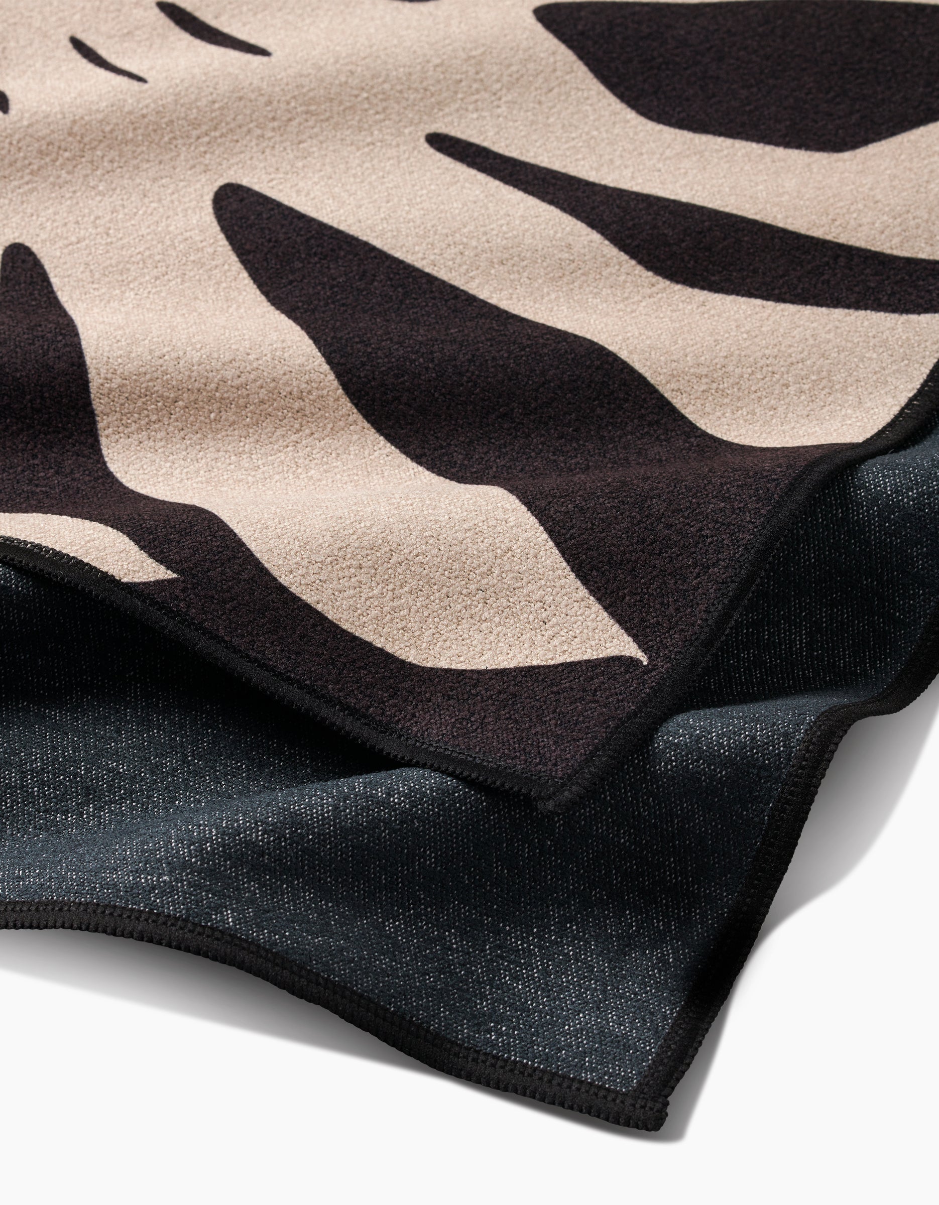 A close-up of two PrintedPlant Fitness Towel fabrics: the Cream Monstera design with bold black shapes over beige on top, and solid black beneath. Both ultra-absorbent towels have finished edges and textured surfaces, shown overlapping flat.