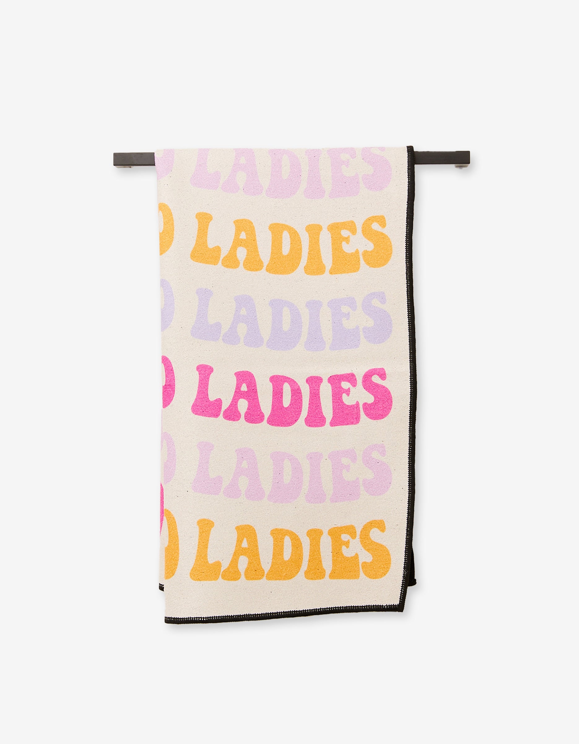The GEOMETRY Ladies Only blanket hangs on a rod, made from recycled materials and showcasing bold pink, orange, and lavender GO LADIES letters on a white background.