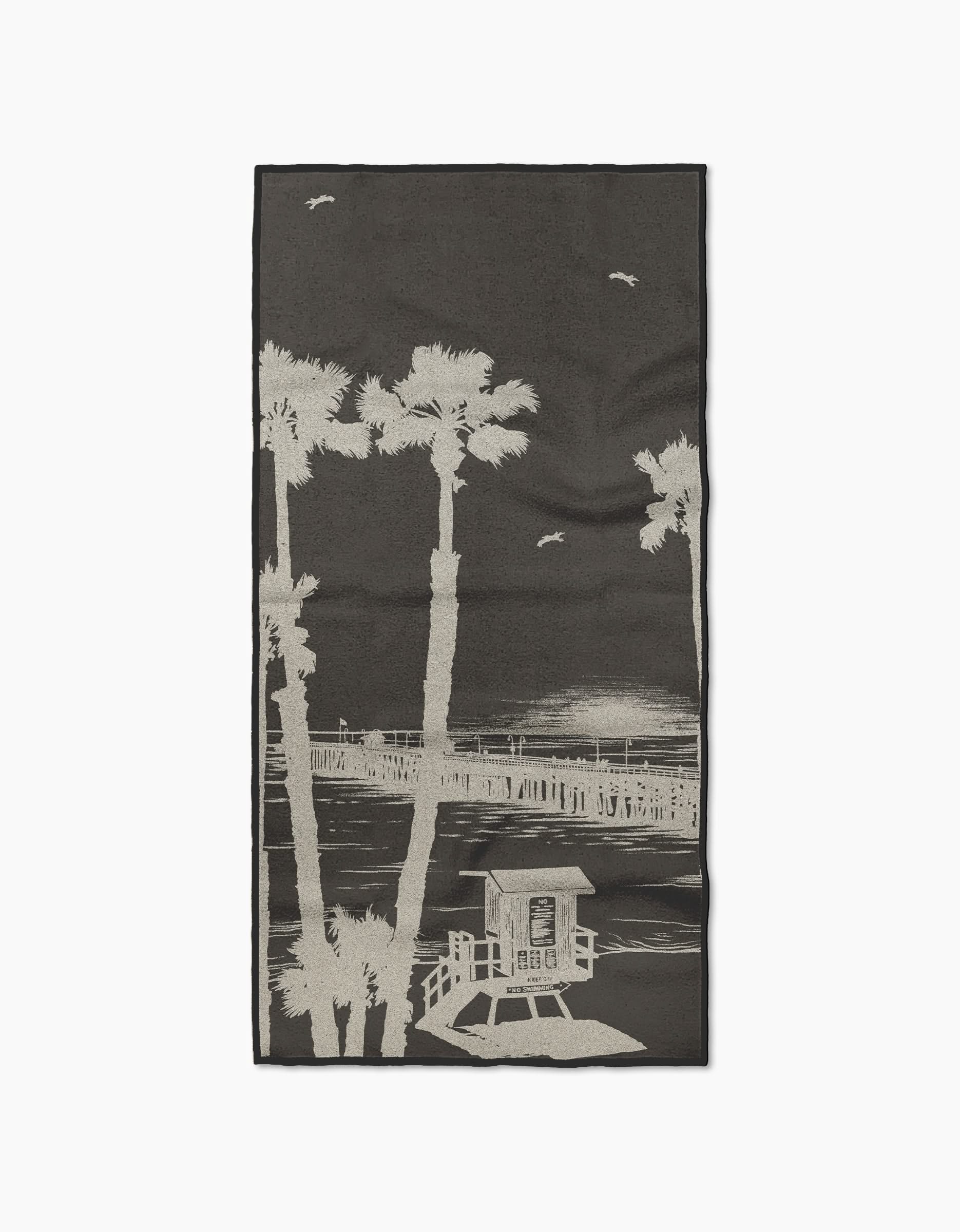 The Geometry Lost In Clemente beach towel features a monochrome palm tree, pier, and lifeguard tower design. Made from recycled materials, it’s fast drying and perfect for comfort at the beach.