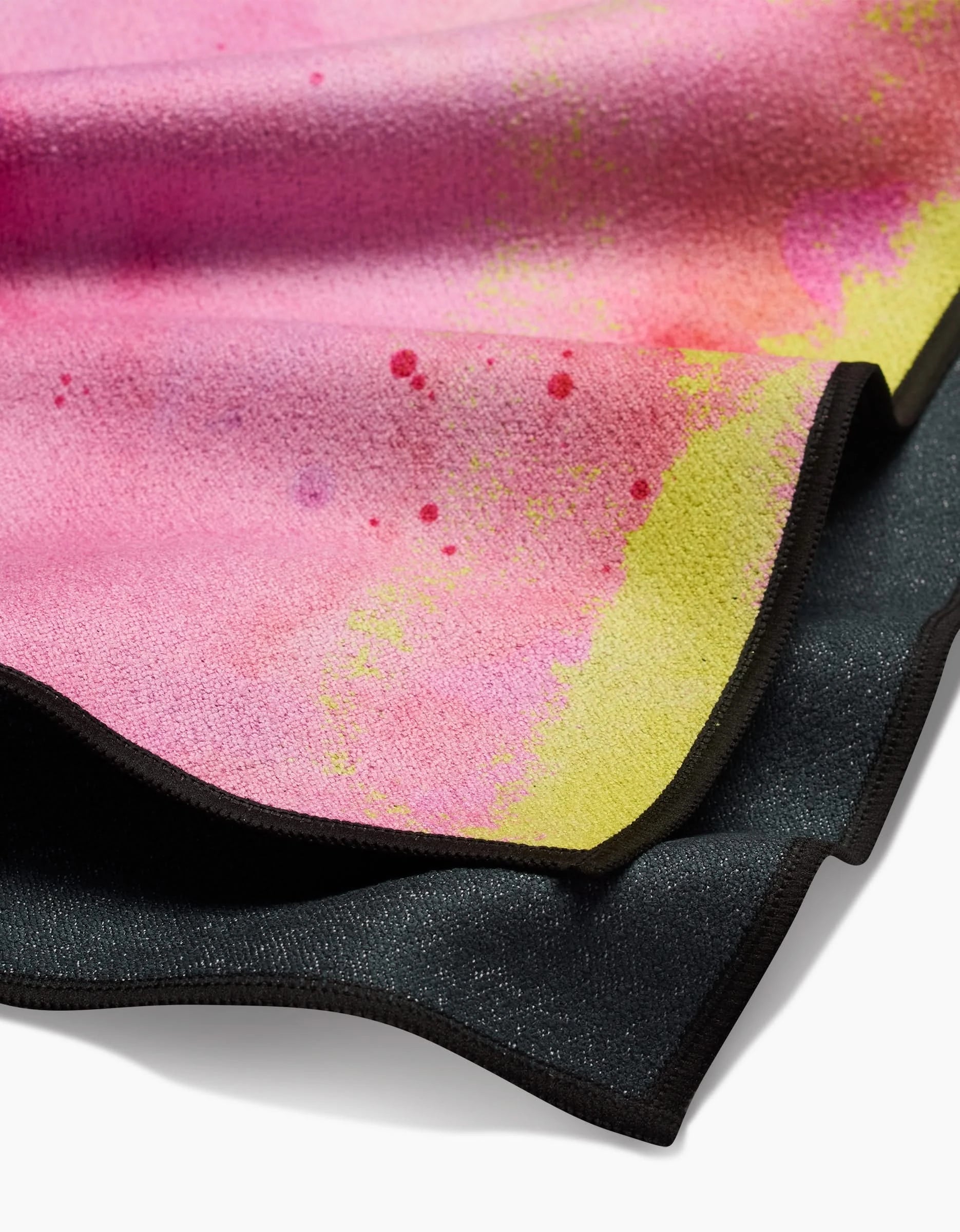 A close-up of GEOMETRY’s Purity towel shows its textured Geoweave fabric with a pink and yellow gradient, splattered paint patterns, and black edging at the bottom.