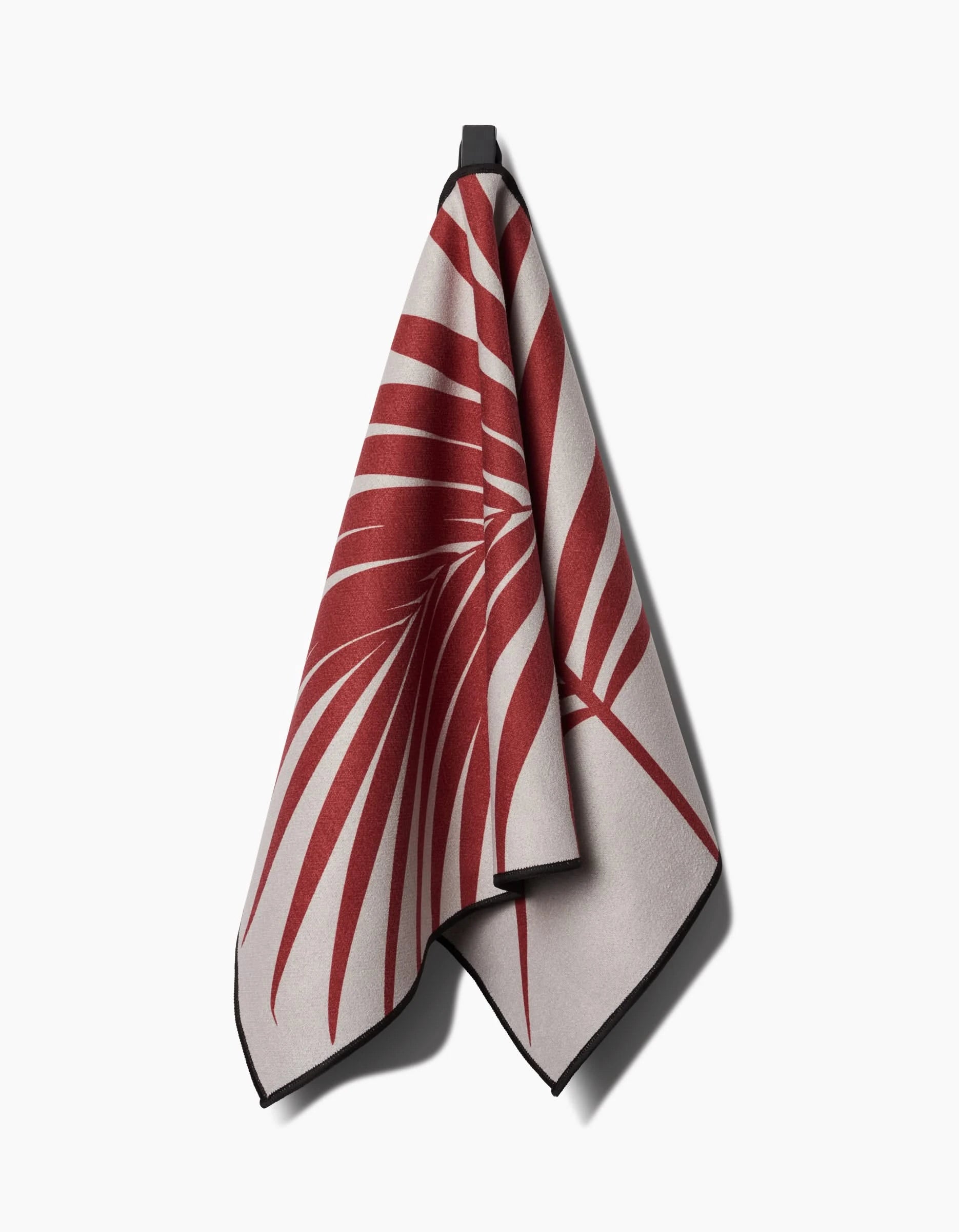 The PrintedPlant Red Palm Fitness Towel, featuring a bold red palm leaf design on beige Geoweave fabric for superior absorbency, hangs from a black hook against a white background.