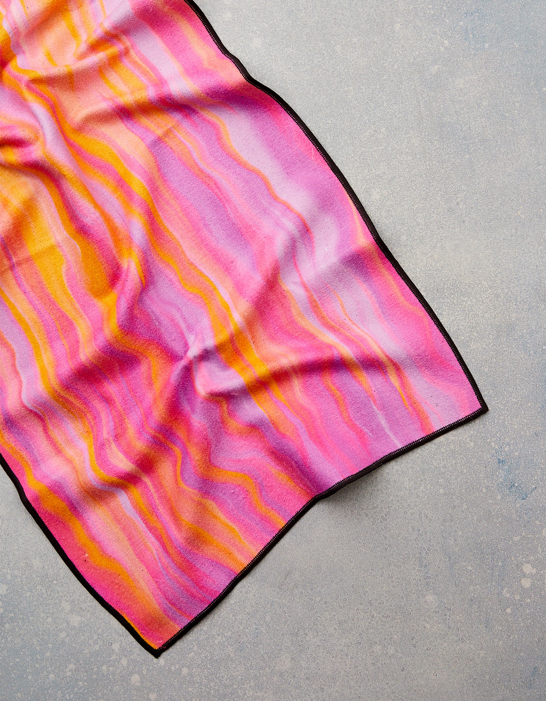 The Rush by GEOMETRY—a vibrant fitness towel with pink, orange, and yellow wavy stripes, thin black trim, and made from recycled materials—is spread out on a light gray textured surface.
