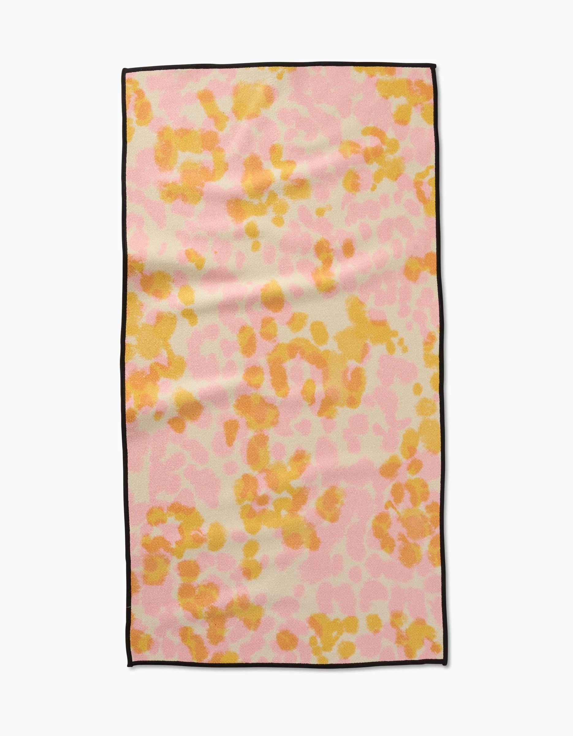 The GEOMETRY Whimsy towel is a rectangular fitness towel made from recycled materials, featuring a pink, orange, and yellow abstract spotted pattern with a thin black edge, shown on a white background.