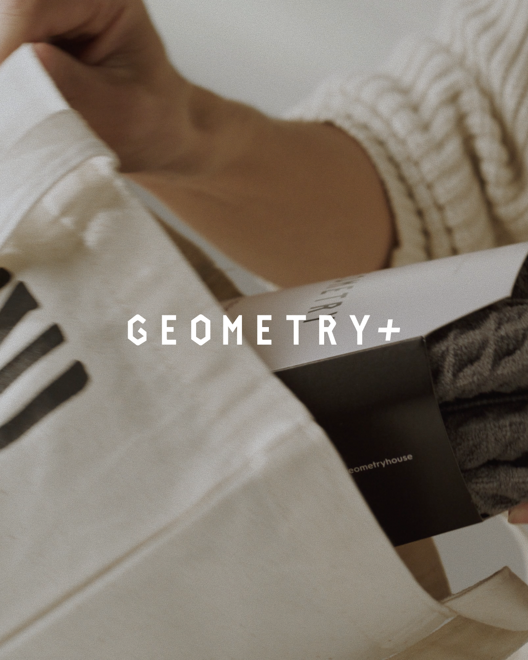A person in a cream knit sweater puts a dark gray, patterned Geometry+ accessory into a white tote bag. GEOMETRY+ appears boldly centered in white over the image, reflecting the modern style of Geometrys latest product.