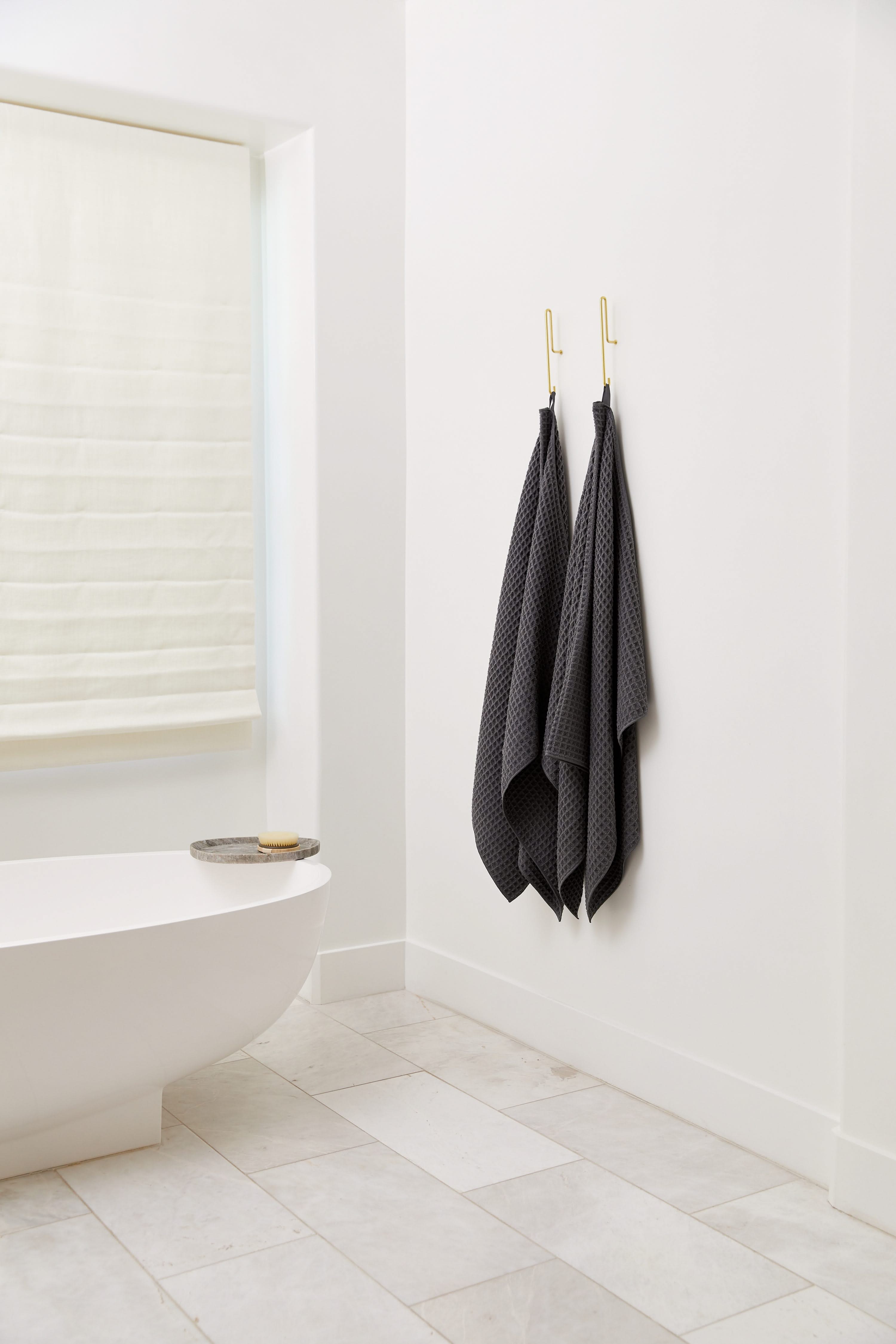 A minimalist bathroom features a white bathtub, light tile flooring, a window with a sheer shade, and two GEOMETRY Charcoal luxury bath towels in dark gray hanging on gold hooks.