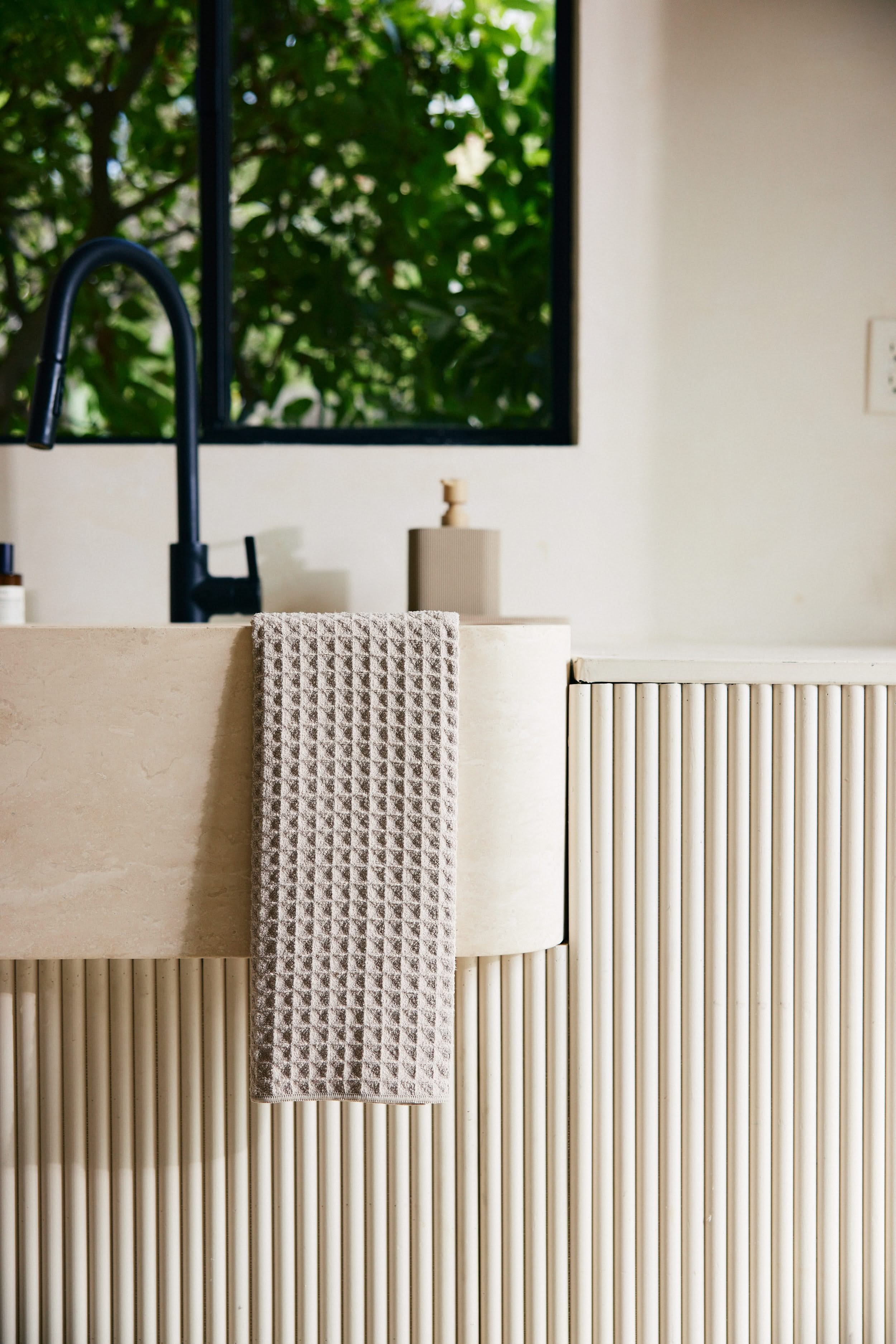 A GEOMETRY Taupe waffle hand towel hangs over a cream-colored, vertically-grooved sink next to a black faucet, with a soap dispenser and a window showing green foliage in the background.