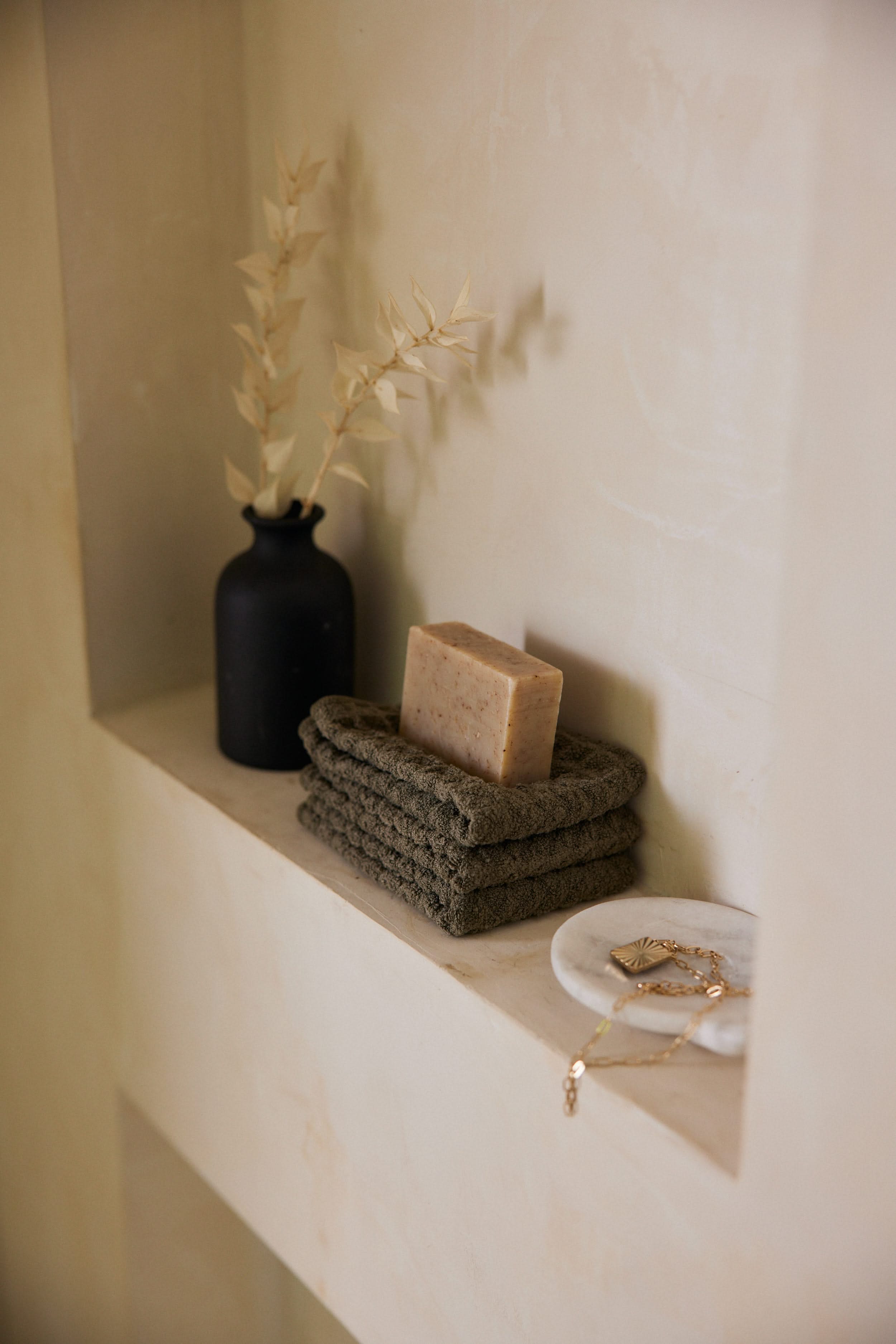 The GEOMETRY Forest bar soap sits atop two plush towels near a black vase with dried beige leaves and a small white dish of gold jewelry, all beautifully arranged on a light-colored shelf.