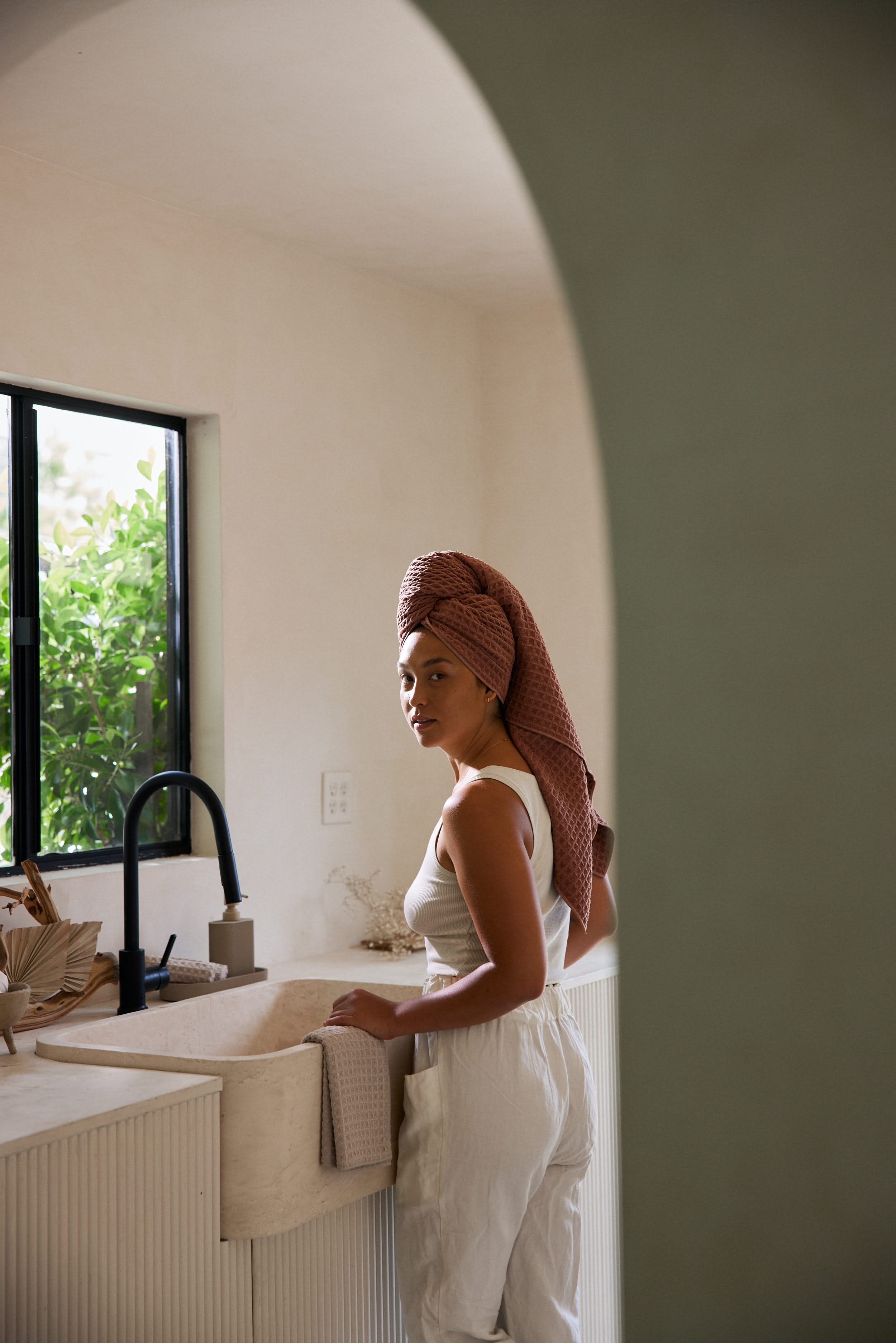 A woman stands at a kitchen sink, wearing a white tank top and pants with a GEOMETRY Dusty Rose luxury towel wrapped around her head. Sunlight streams through the window, revealing greenery outside as she holds a cloth.