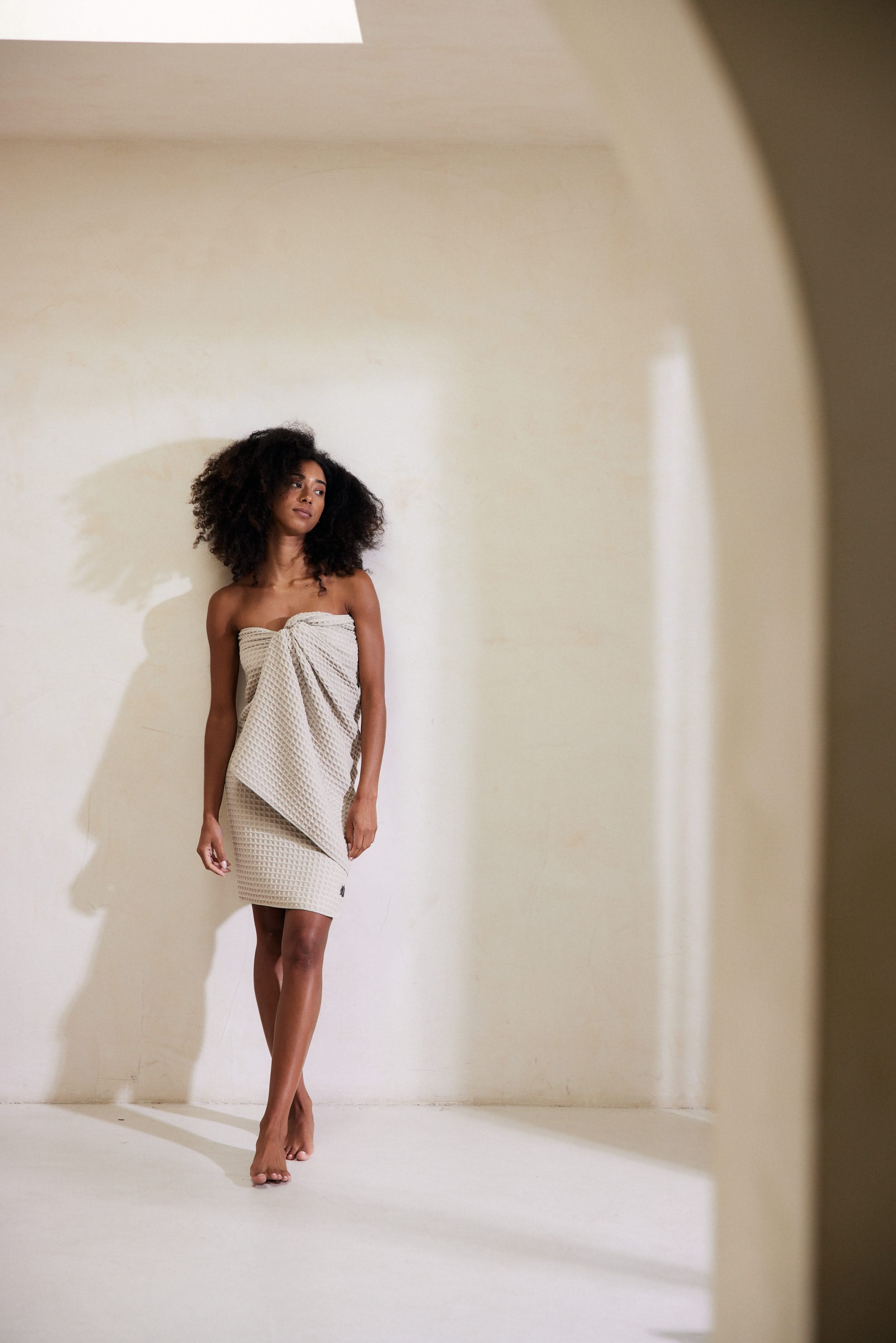 A woman with natural curls stands barefoot in a bright, minimalist room, wearing GEOMETRYs Taupe, a strapless textured cream dress made from recycled materials. She gazes to the side, casting a gentle shadow on the wall.