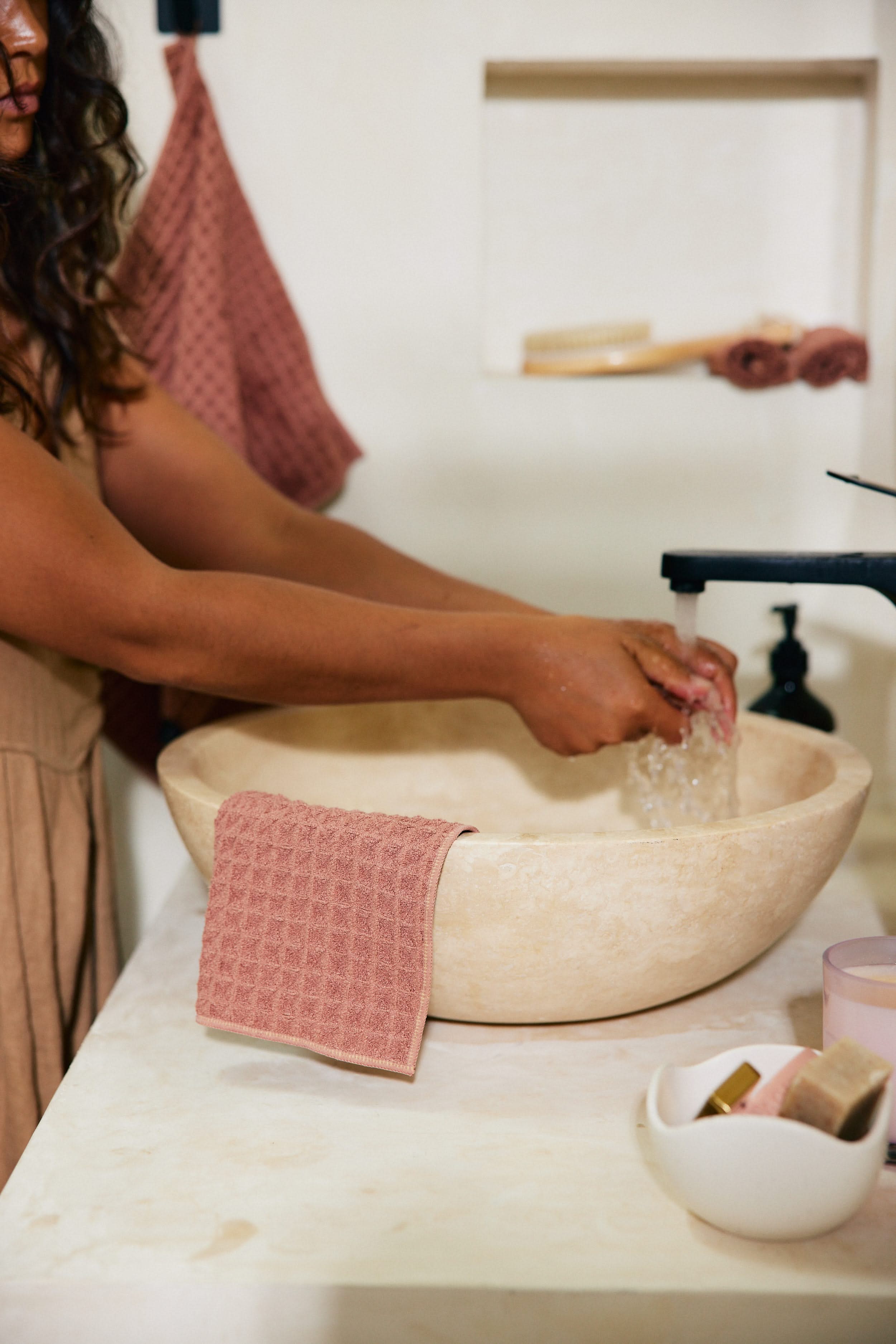 A person washes their hands at a stone sink with a Dusty Rose towel by GEOMETRY draped over the edge. Matching pink and beige GEOMETRY bath accessories and towels are seen in the background.
