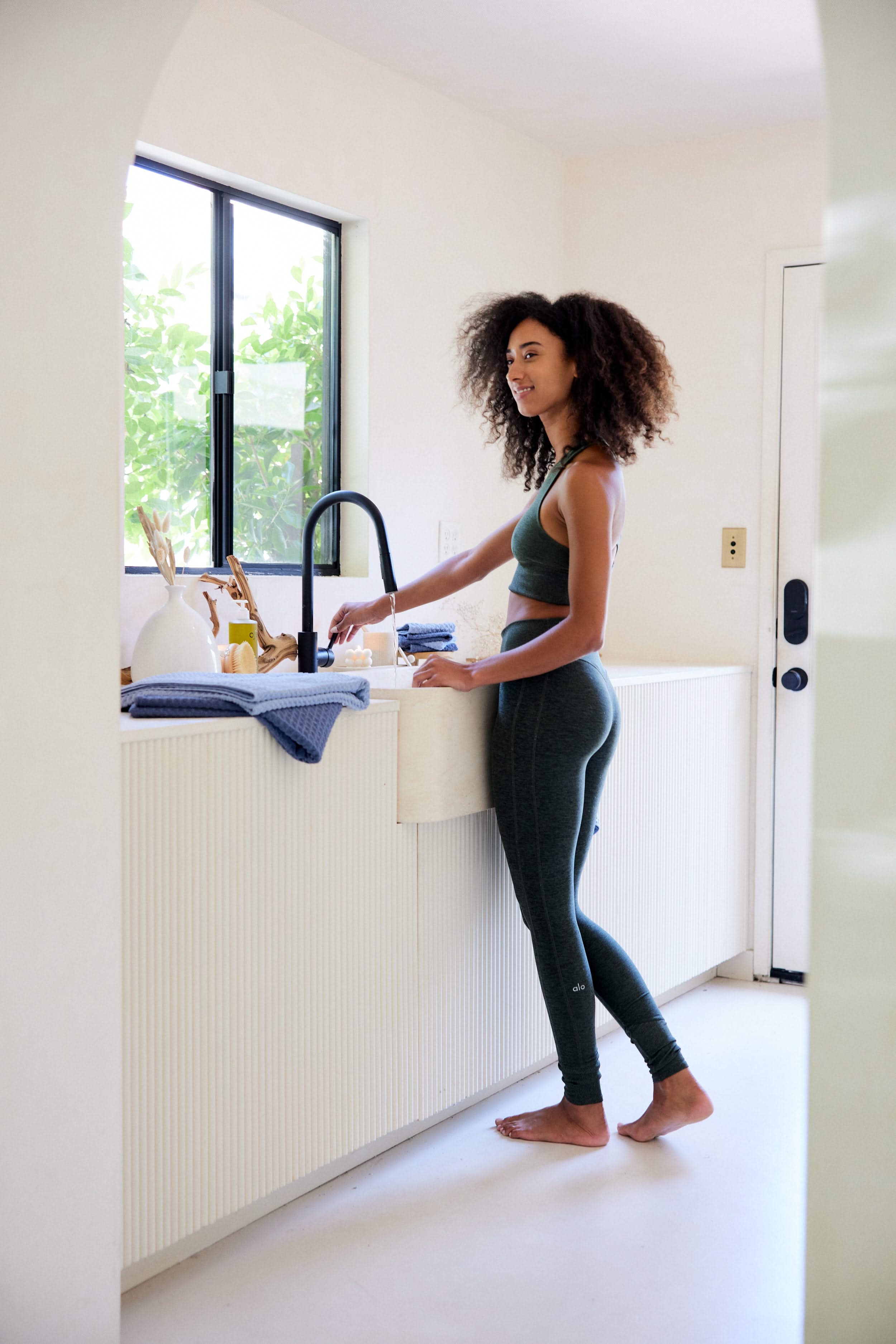 A woman in green activewear smiles as she washes dishes barefoot at a sunlit kitchen sink. On the counter are eco-friendly GEOMETRY towels in Midnight Blue, along with sustainable utensils. Green foliage is visible through the window.