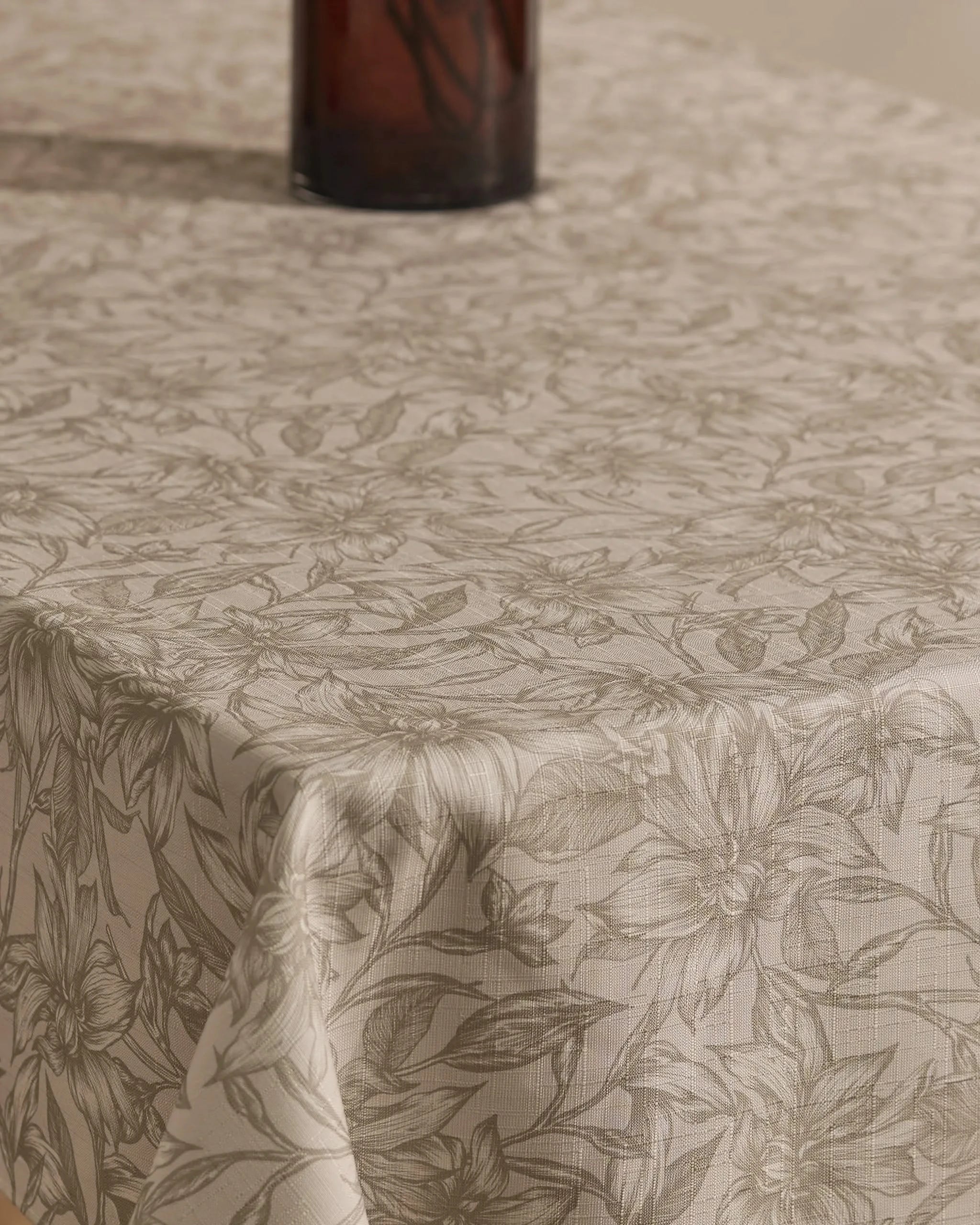 A close-up of the Geometry Alba tablecloth, beige and stain-resistant with a subtle floral pattern, covers the table. The bottom of a dark glass vase appears at the images top.