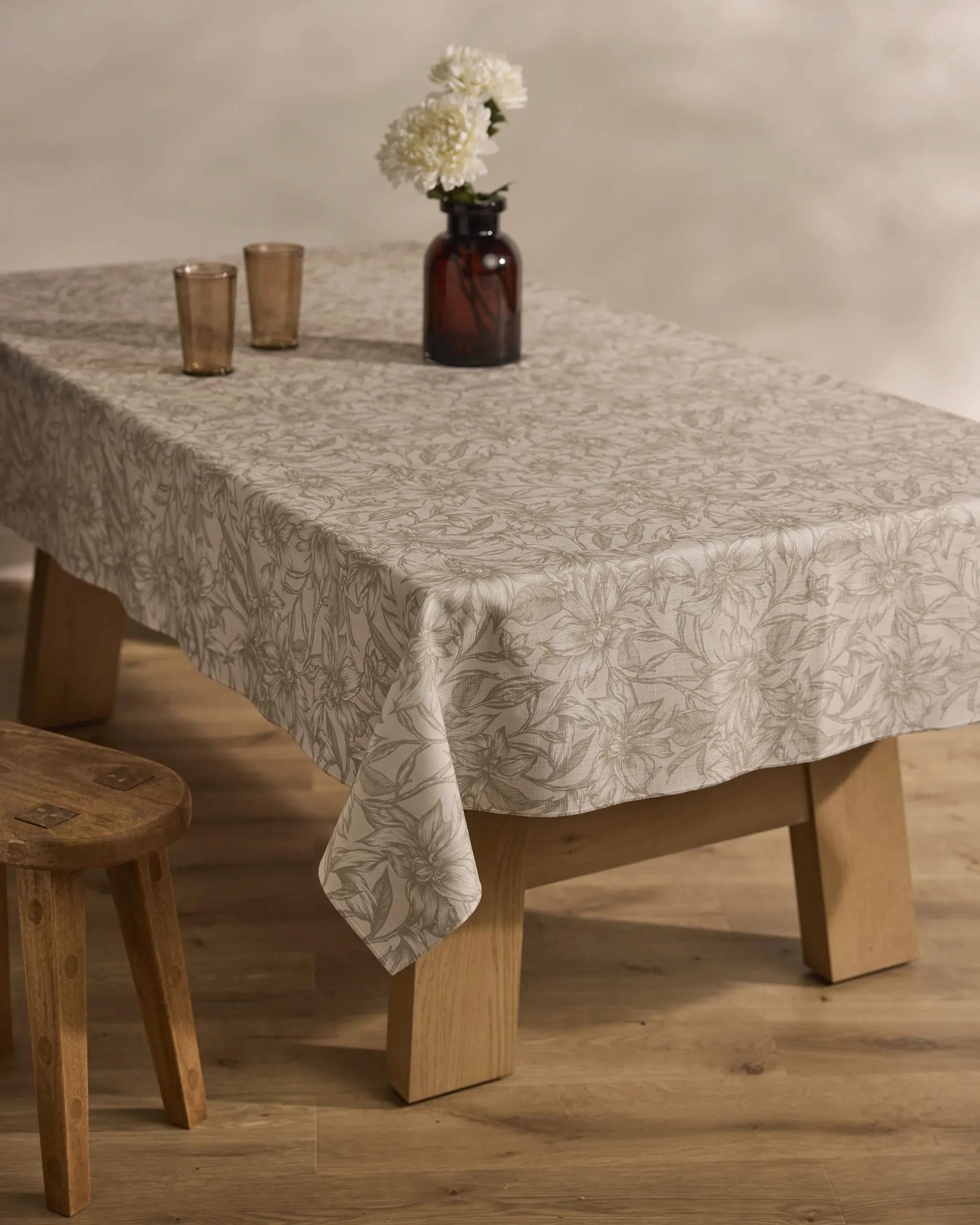 The Alba table by Geometry features a floral-patterned, stain-resistant tablecloth, two brown glasses, and a dark vase with white flowers. A wooden stool is nearby, all set on a light wood floor.