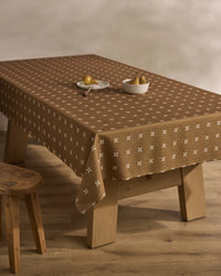 The Amber Stitch tablecloth by Geometry, brown and stain-resistant with a white cross pattern, covers a wooden table set with pears and a bowl. A wooden stool stands nearby on a light wood floor.