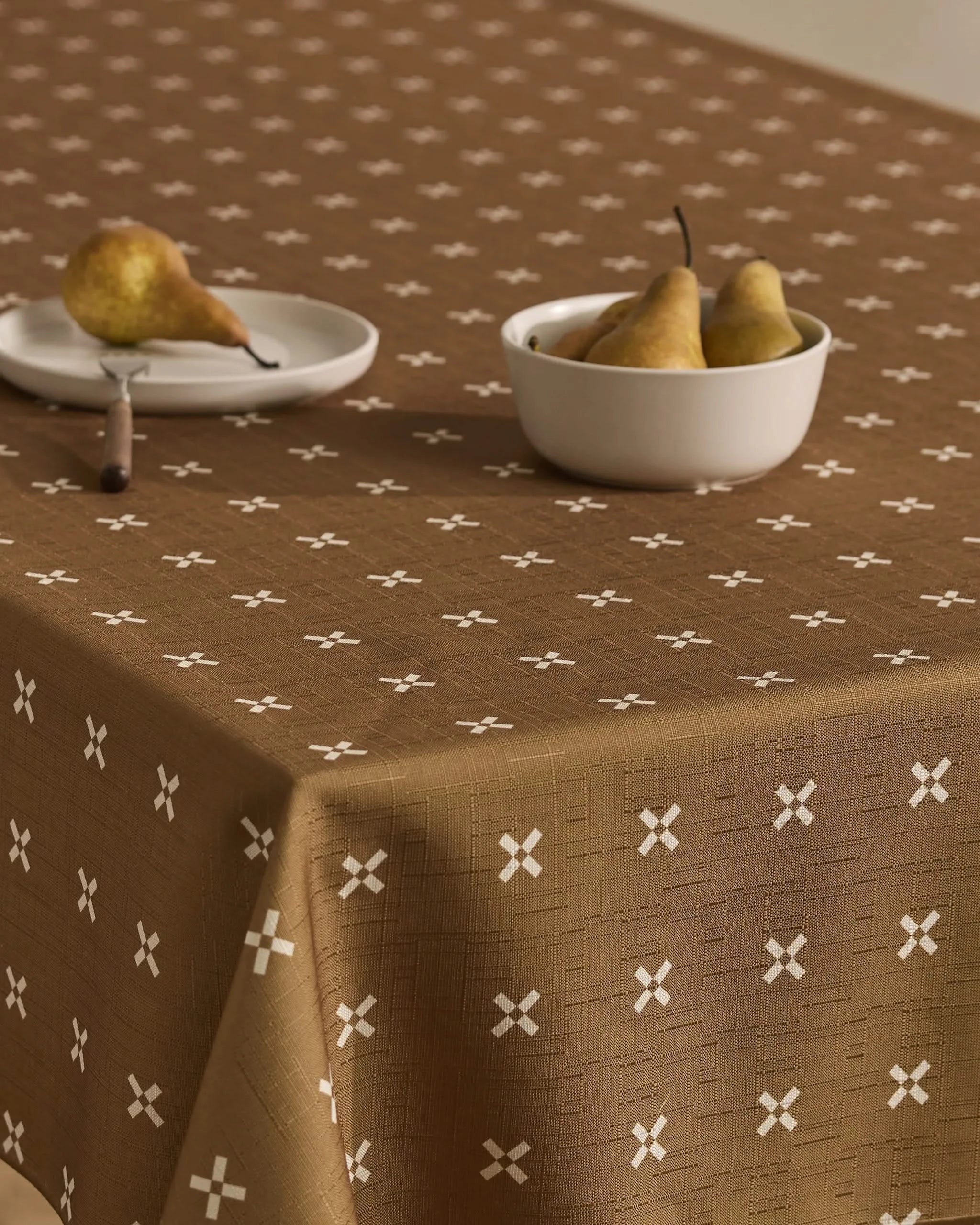 The table is covered with the Amber Stitch tablecloth by Geometry, featuring a white X pattern. A white plate with a pear and a bowl with three pears sit on top, set against a neutral, minimal background.