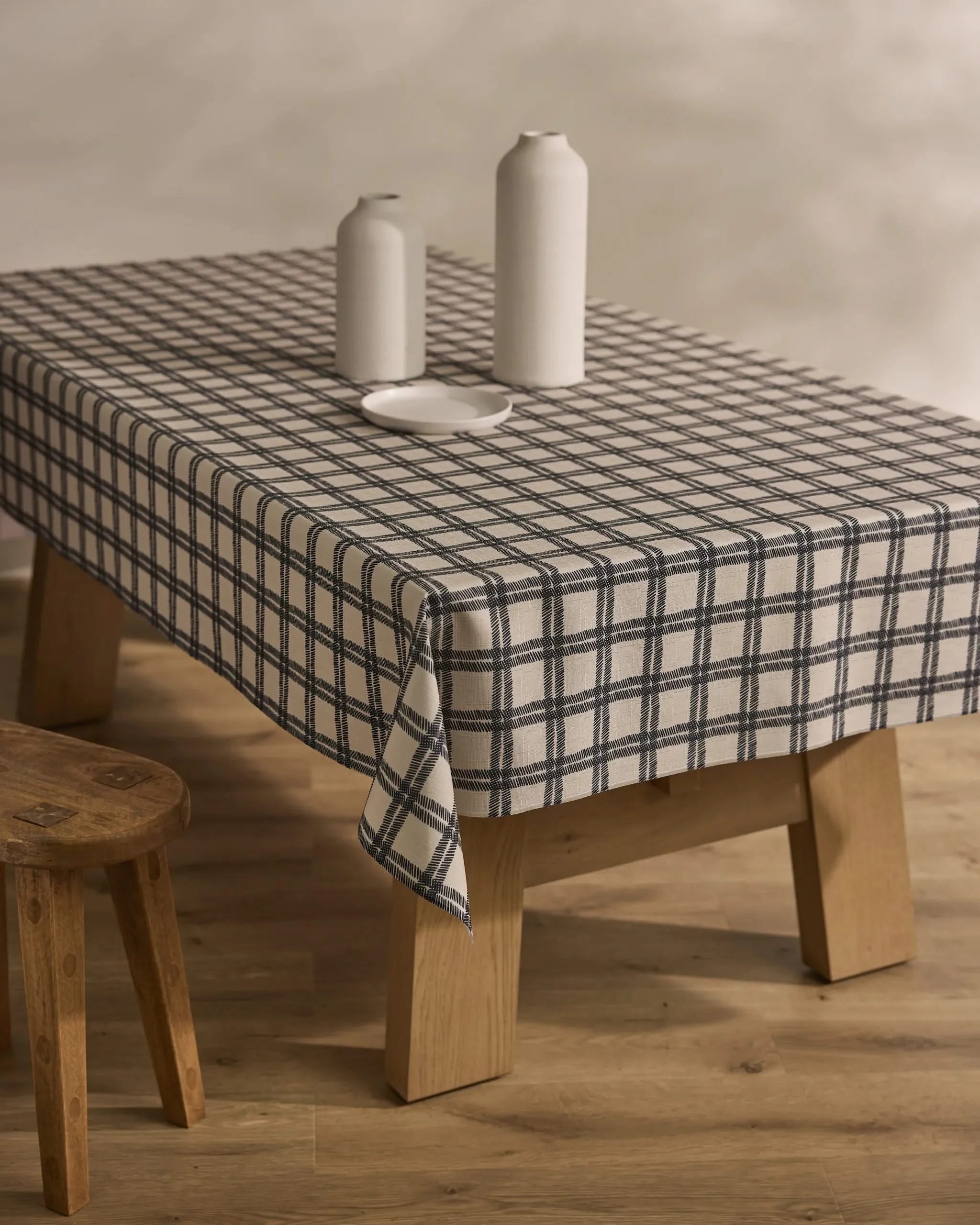 The Blackstone table by Geometry, topped with a stain-resistant white and blue checkered cloth, displays three tall white vases and a small white plate. A wooden stool stands nearby on the wooden floor.