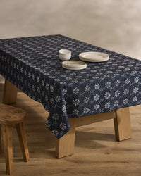 The Bonnie Indigo tablecloth by Geometry, featuring a white floral pattern on dark blue, covers a wooden table set with three stacked white plates and two white bowls. A wooden stool sits beside the table on a light wood floor.