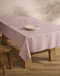 A wooden table draped with the Geometry Bubble Ditzy Dusty Rose stain-resistant tablecloth, featuring subtle patterns, is set with three stacked white plates and two clear glasses beside a basic wooden stool on a wood floor.