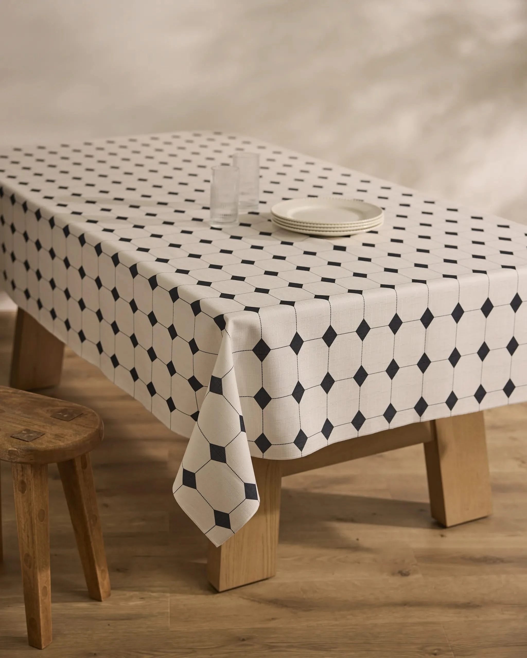 The Espresso Mosaic by Geometry is a rectangular wooden table draped with a lightweight white cloth featuring a black geometric pattern. Two clear glasses and stacked white plates sit on top, with a wooden stool beside it on a light wood floor.
