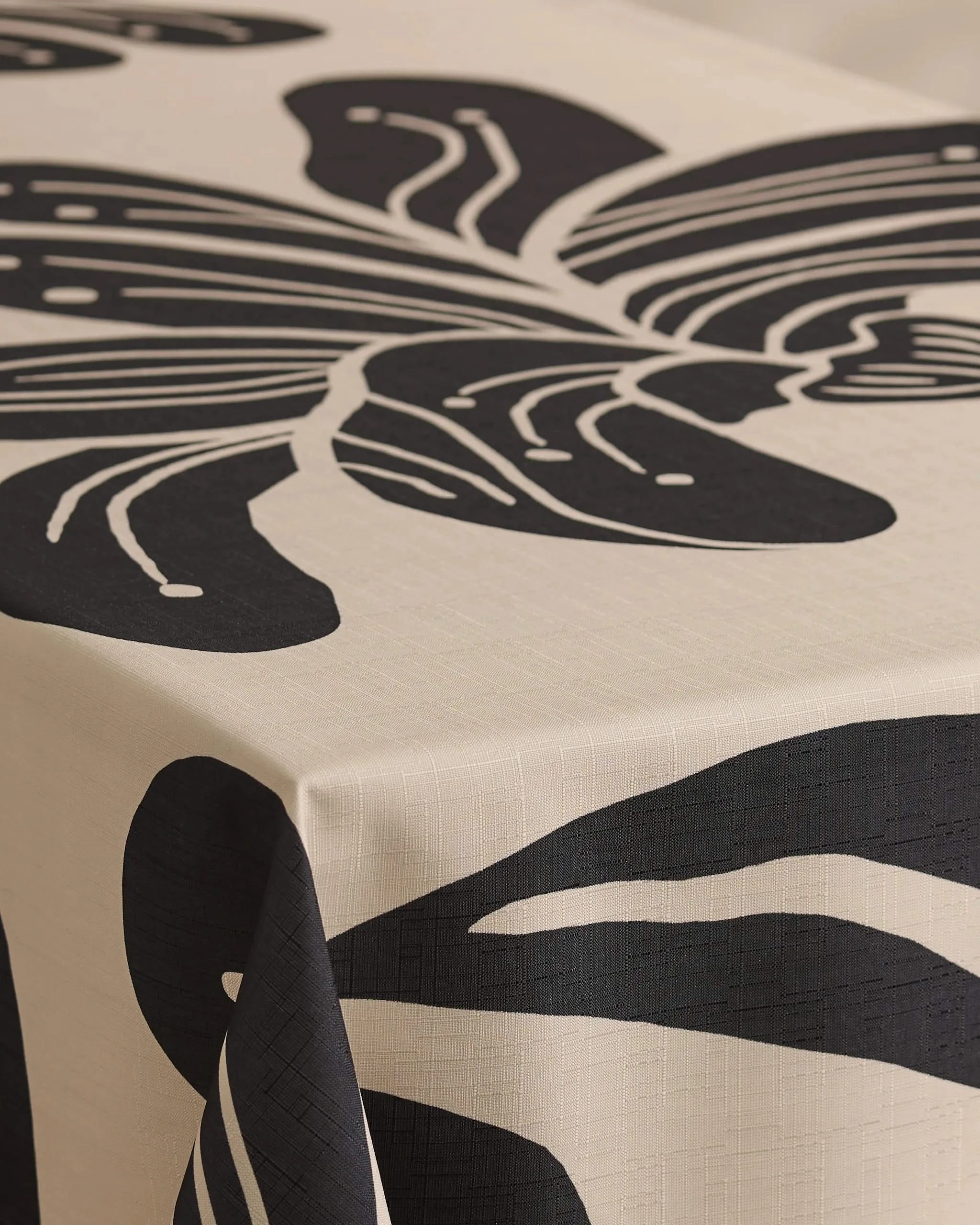 A close-up of a table featuring Geometrys Giant Lush Floral tablecloth, cream-colored and stain-resistant, decorated with a bold abstract black leaf design that extends over the edge.
