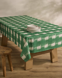 A wooden table is draped with the Geometry Hairy Check Green tablecloth and set with stacked white plates. A wooden stool sits nearby on a light wood floor, all lit by soft, natural light.