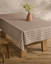 The Hazel Gingham tablecloth by Geometry, in lightweight beige and white checks, covers a wooden table topped with a brown jug of green foliage, beside a wooden stool on a light wood floor.