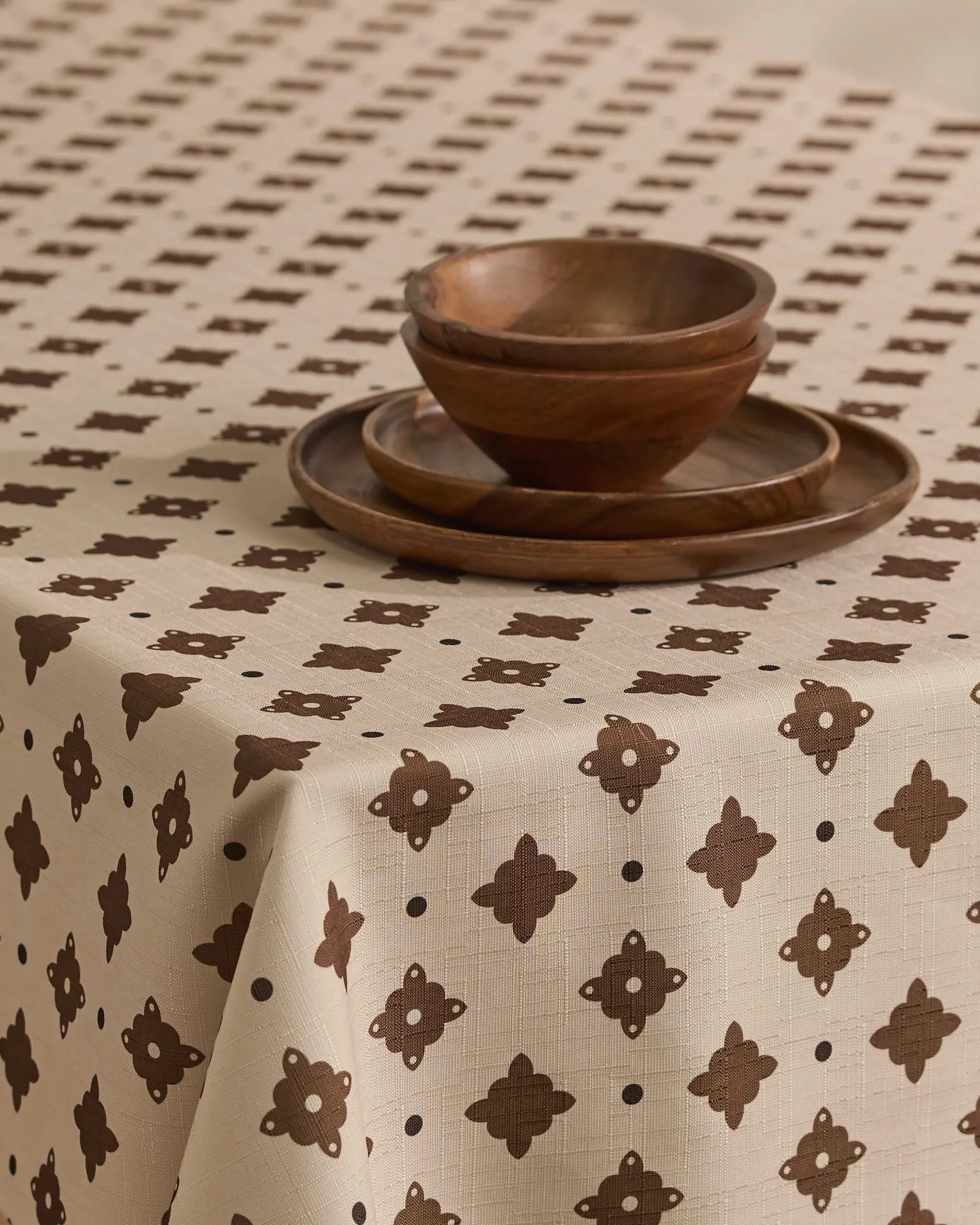 The Heritage Stamp tablecloth by Geometry, in beige with a brown geometric pattern, covers the table. Two wooden plates are stacked and two wooden bowls are nested, creating a simple, rustic setting.